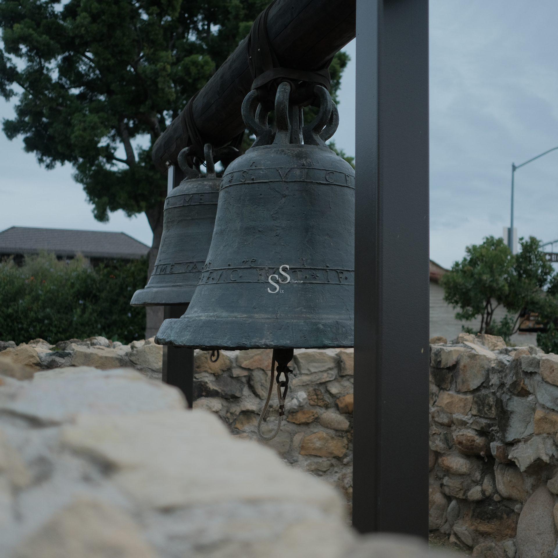 Two large bronze mission bells hang from a wooden beam supported by metal posts, surrounded by a stone wall, with trees and buildings visible in the background.