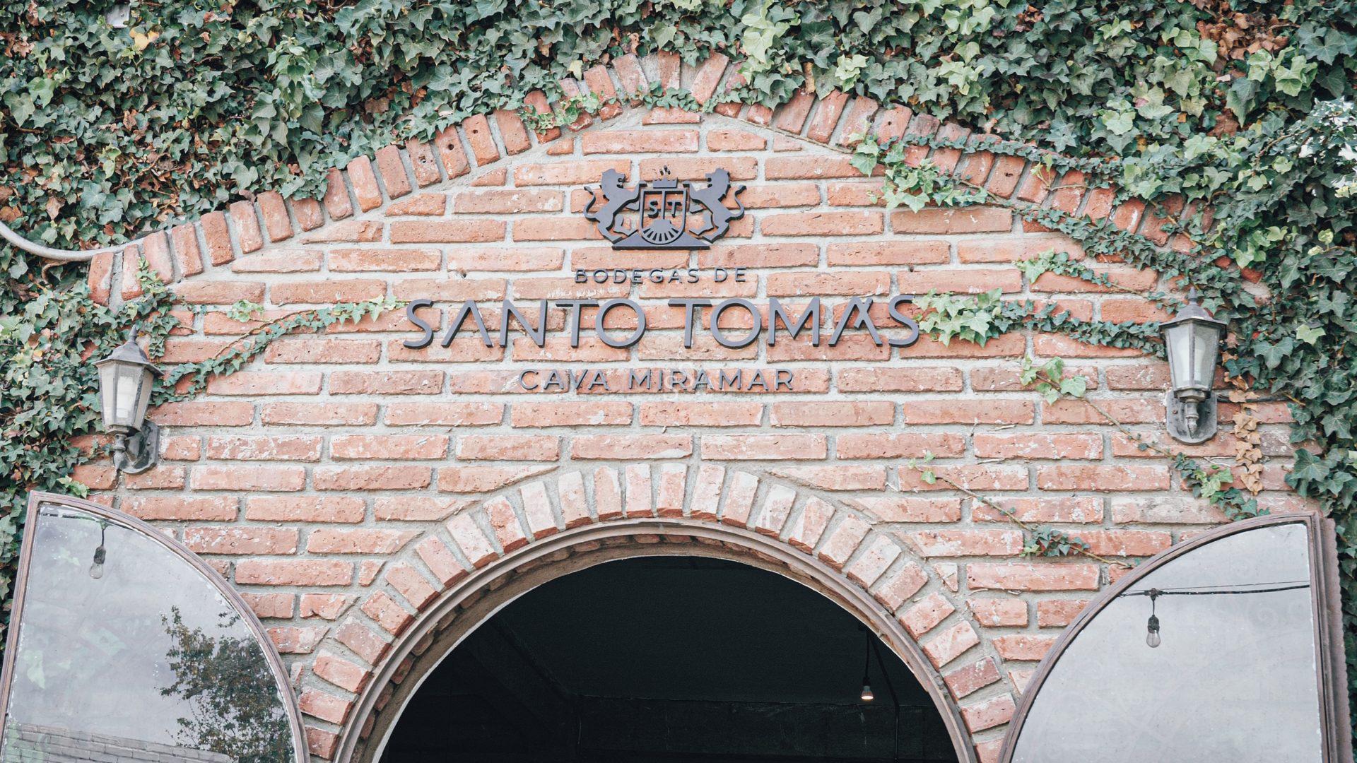 A brick archway covered with green ivy, featuring the sign Bodegas de Santo Tomás Cava Miramar and two wall-mounted lanterns on either side. Glass doors are partially open below the sign.