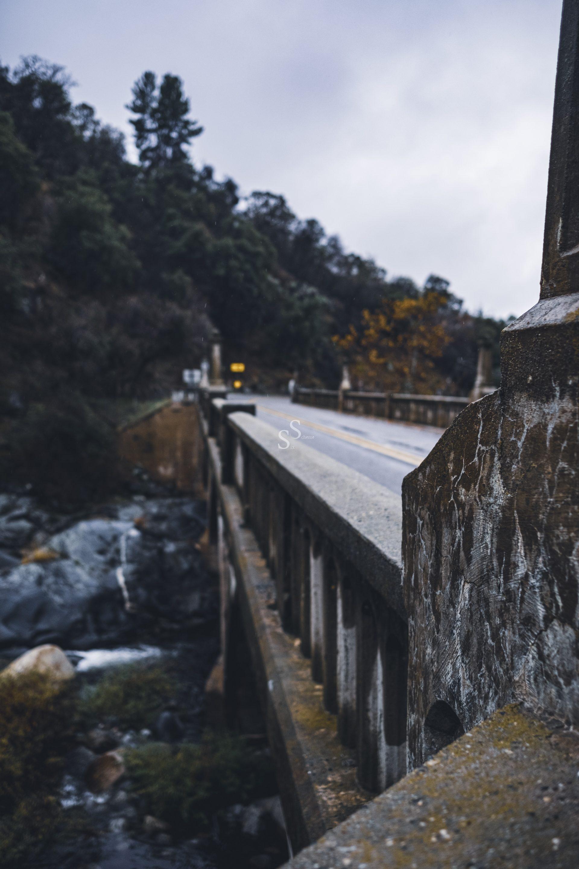 A stone bridge with weathered railings stretches over a rocky stream, surrounded by trees and hills under a cloudy, overcast sky. The scene appears calm and somewhat moody.