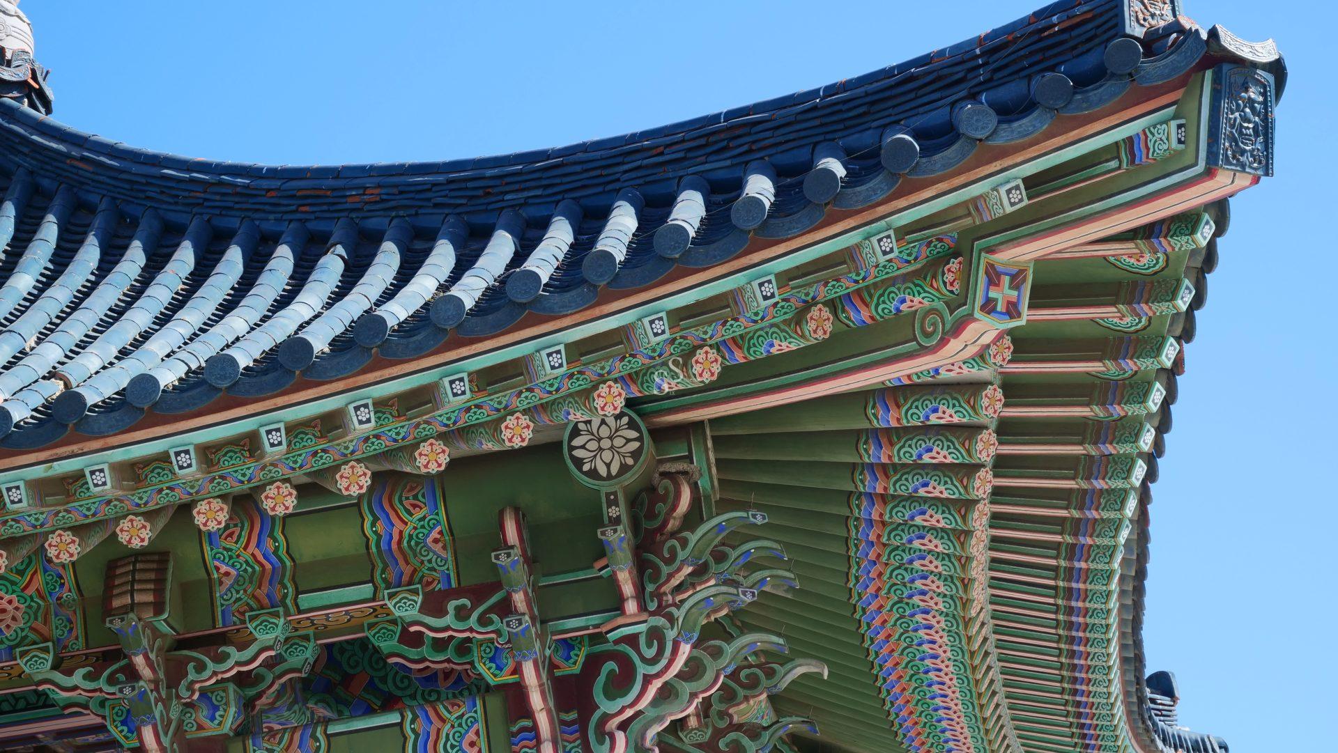 A close-up of the ornate, colorful eaves of a traditional Korean building, featuring intricate painted patterns and wooden carvings, set against a clear blue sky.