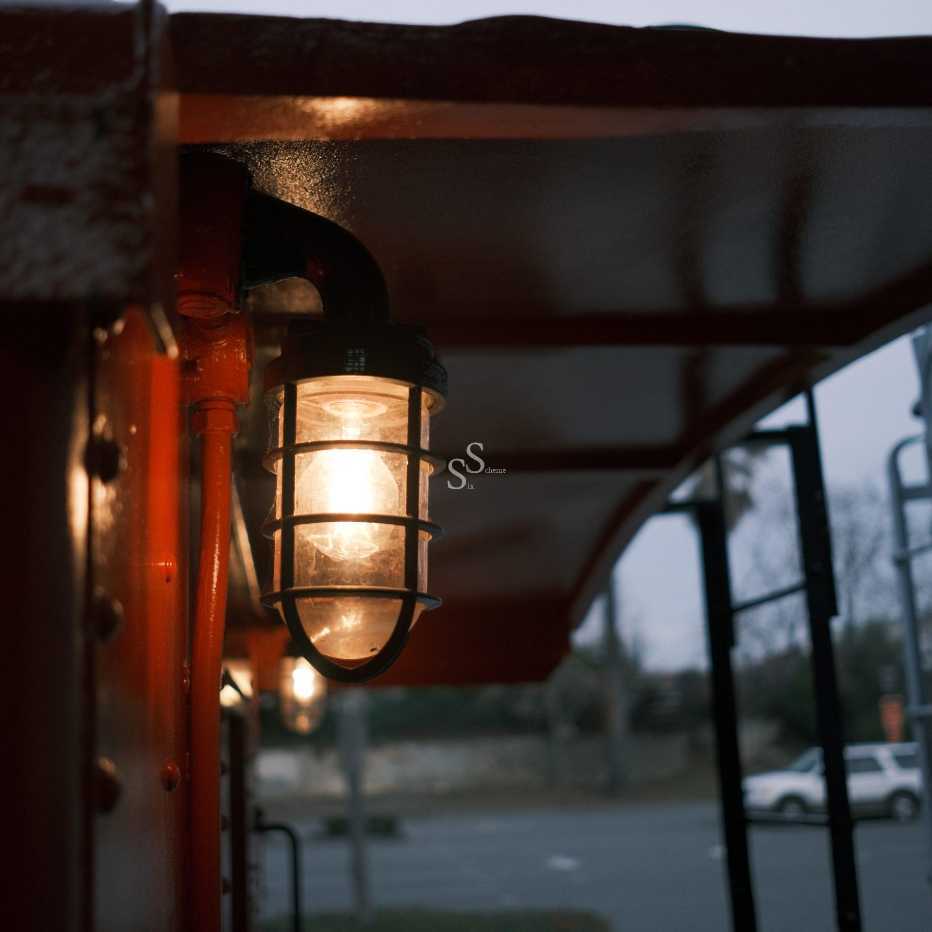 A close-up of a lit industrial-style lamp with a metal cage, mounted on the red exterior of a vehicle or structure, with a blurred street and parked car visible in the background.