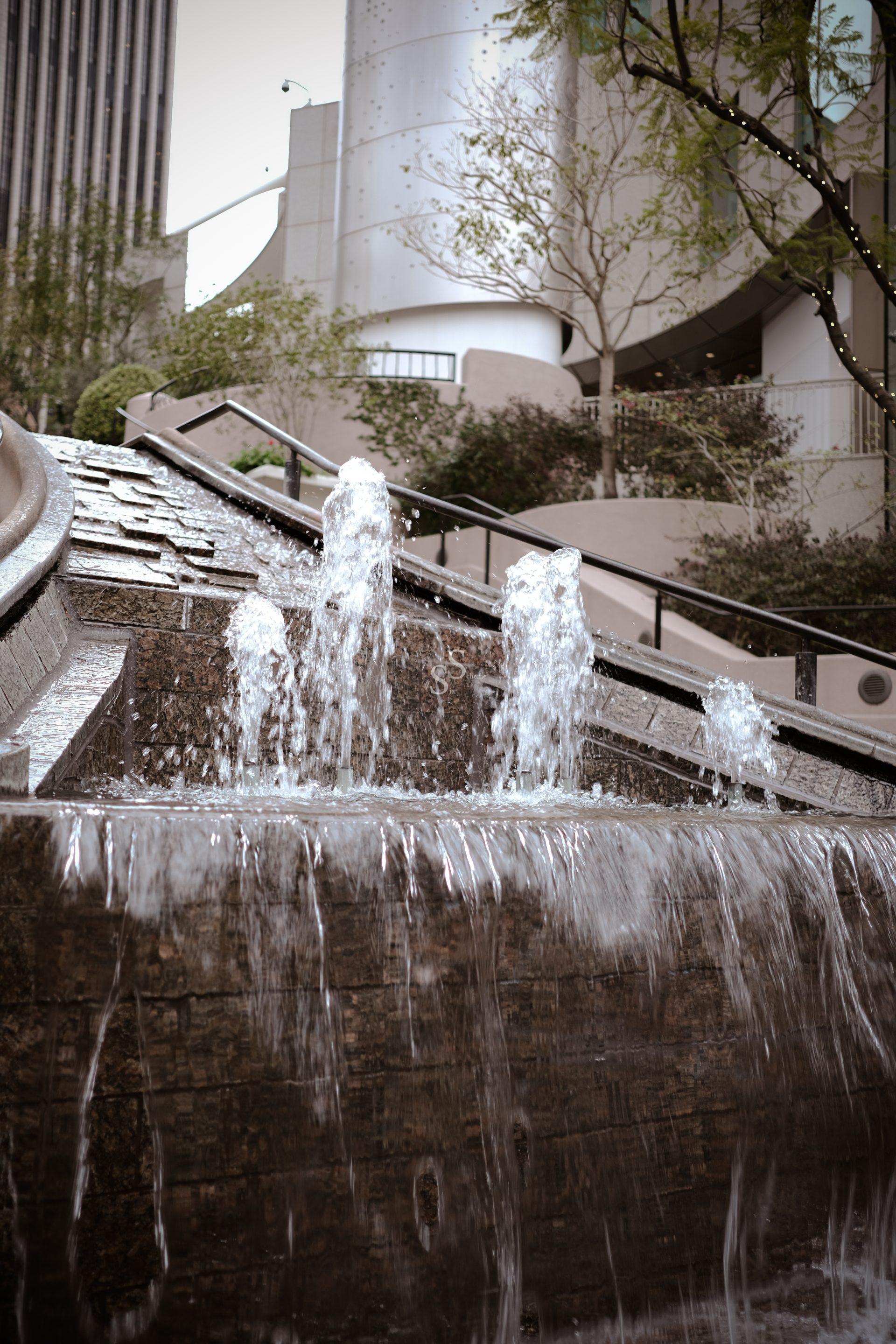 A close-up of a modern urban fountain with water jets and cascading streams, set against a backdrop of city buildings, trees, and stairs with railings.