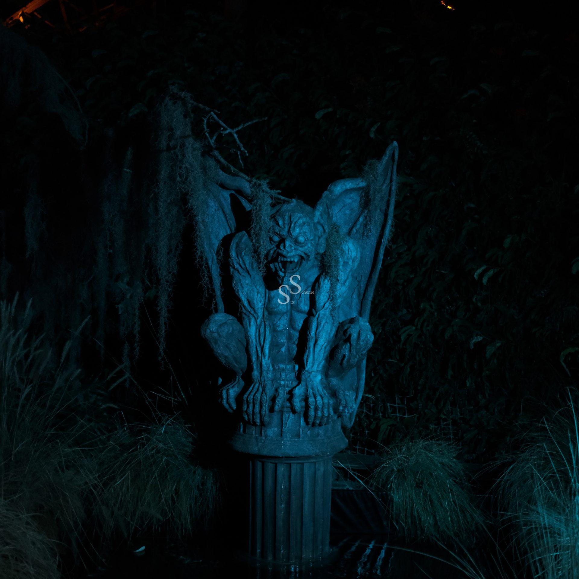 A menacing gargoyle statue with wings crouches on a pedestal, illuminated by eerie blue light and surrounded by tall grass in a dark, shadowy setting.