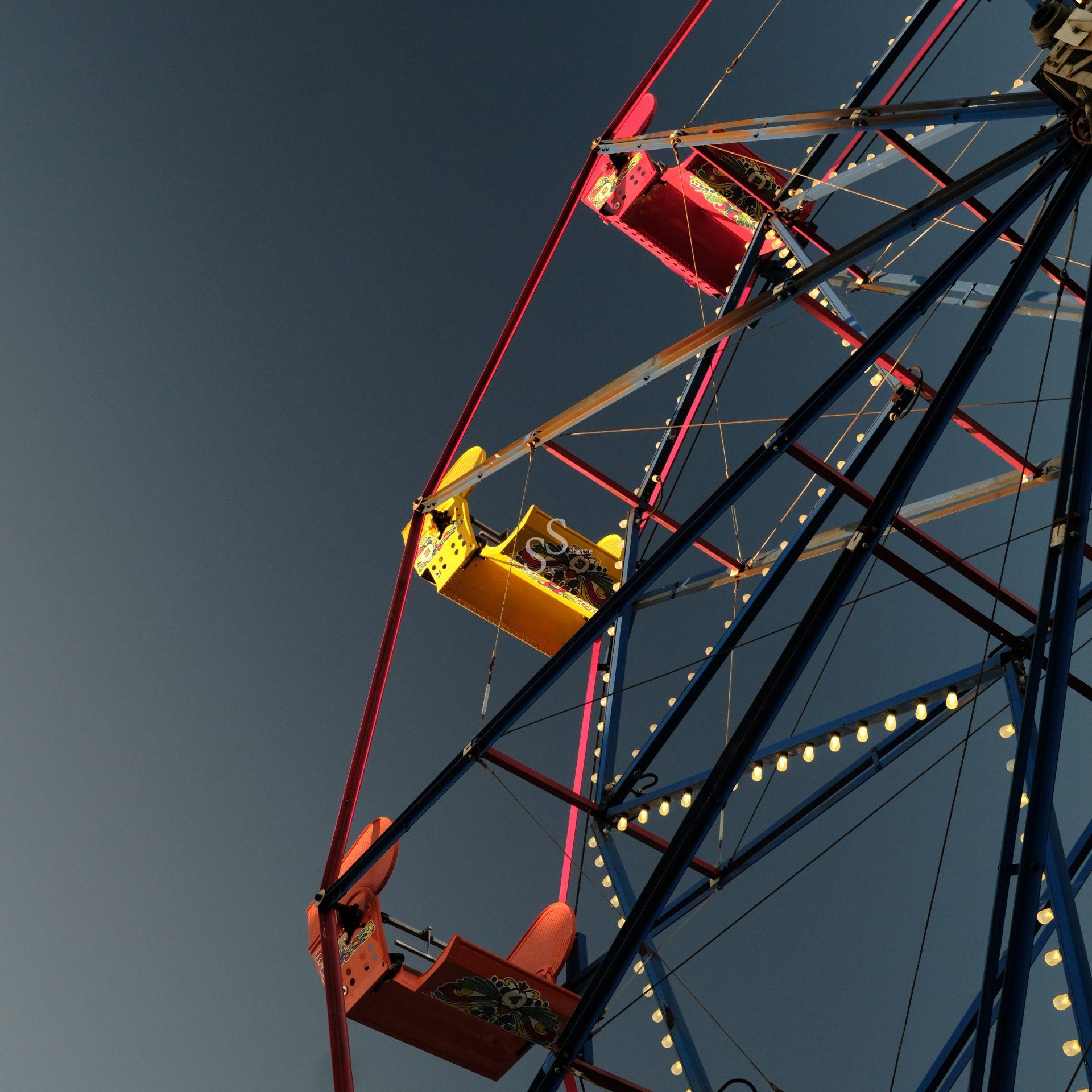 A close-up view of a Ferris wheel with red, yellow, and pink gondolas against a clear, deep blue sky, taken from a low angle. The structure is lit by small lights along its frame.