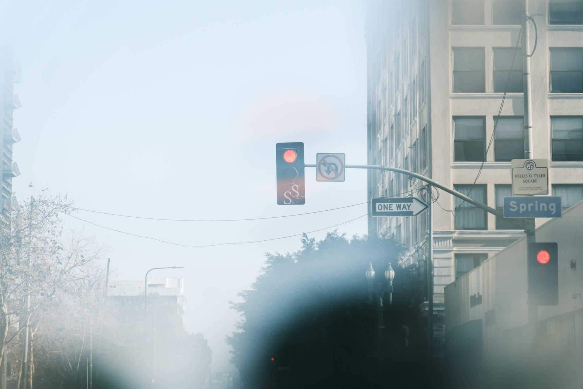 A red traffic light at an intersection with a One Way and Spring street sign, surrounded by tall buildings and some tree branches, viewed through a foggy or blurry lens.