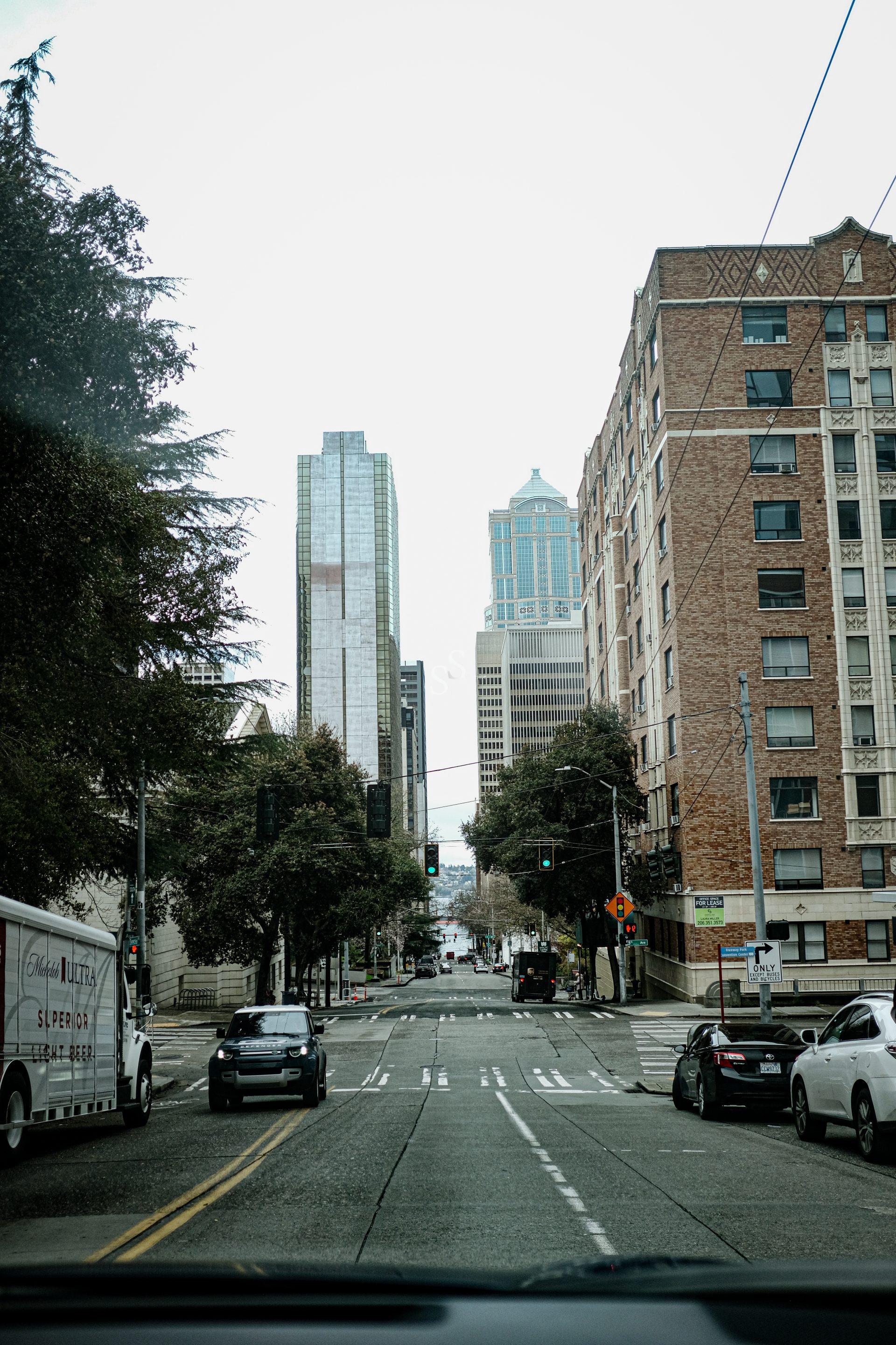 A city street view with cars driving and parked, tall buildings in the background, trees lining the road, and traffic lights overhead under a cloudy sky.