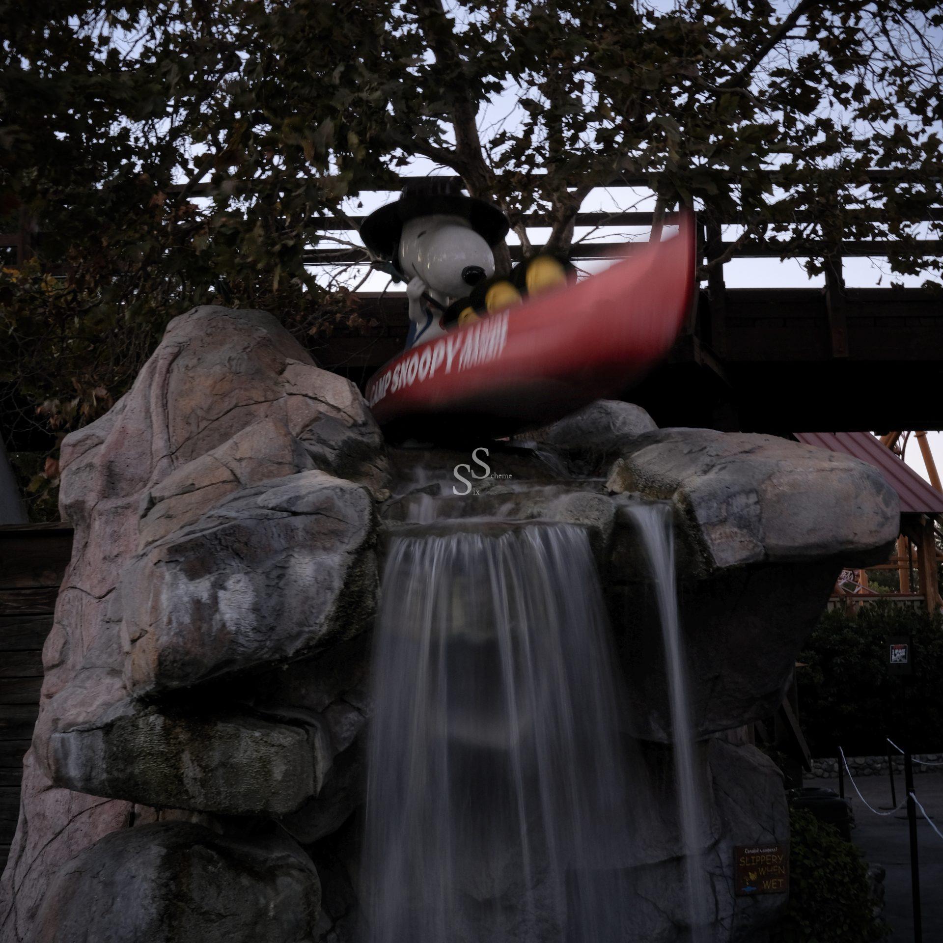 A Snoopy statue wearing a hat sits in a red canoe atop artificial rocks with water flowing down like a waterfall, surrounded by trees and a bridge overhead.