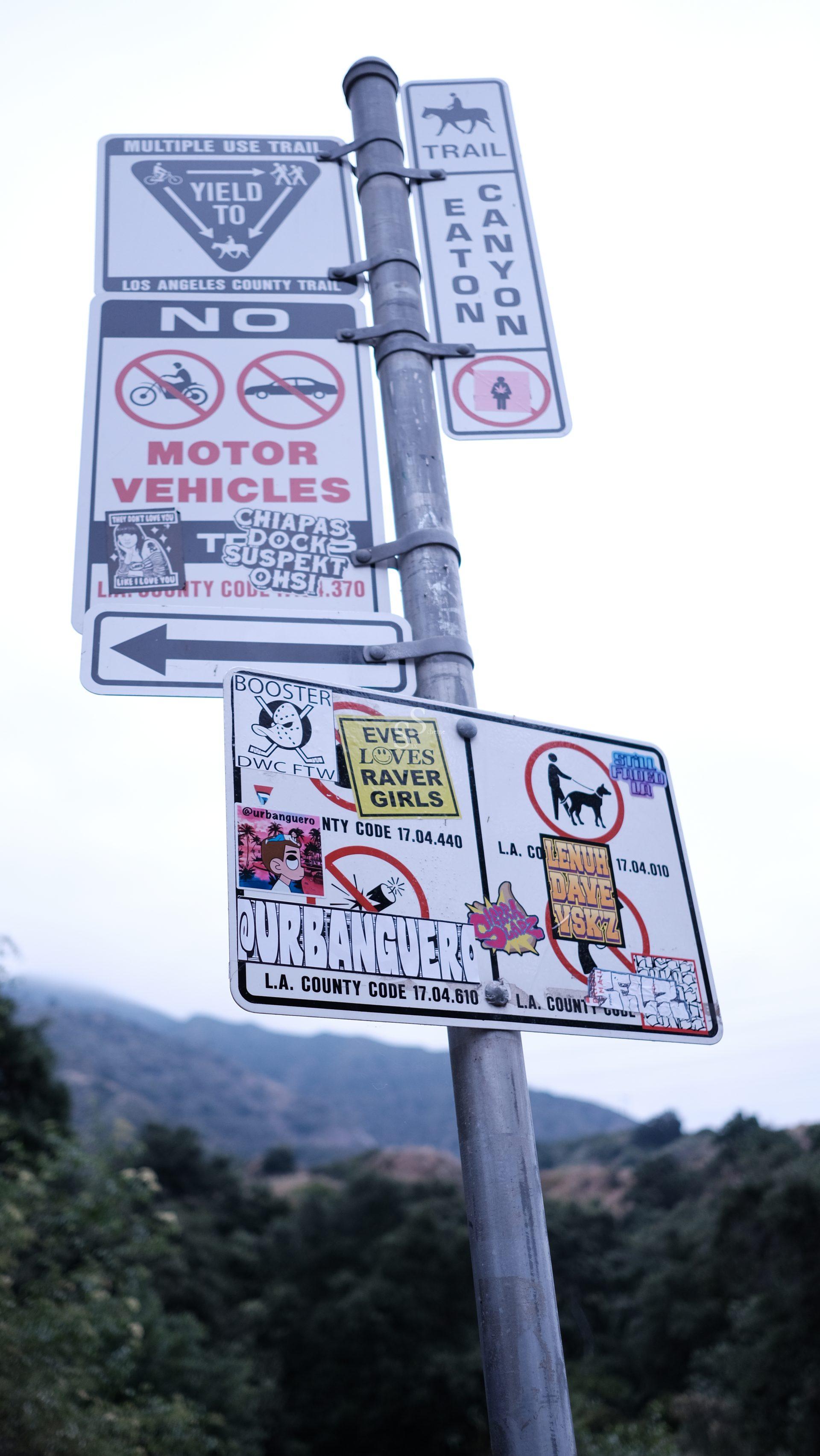 A metal pole with multiple trail and warning signs, including “Yield to Hikers,” “No Motor Vehicles,” and “Eaton Canyon Trail.” Signs are covered with various colorful stickers; mountains are visible in the background.