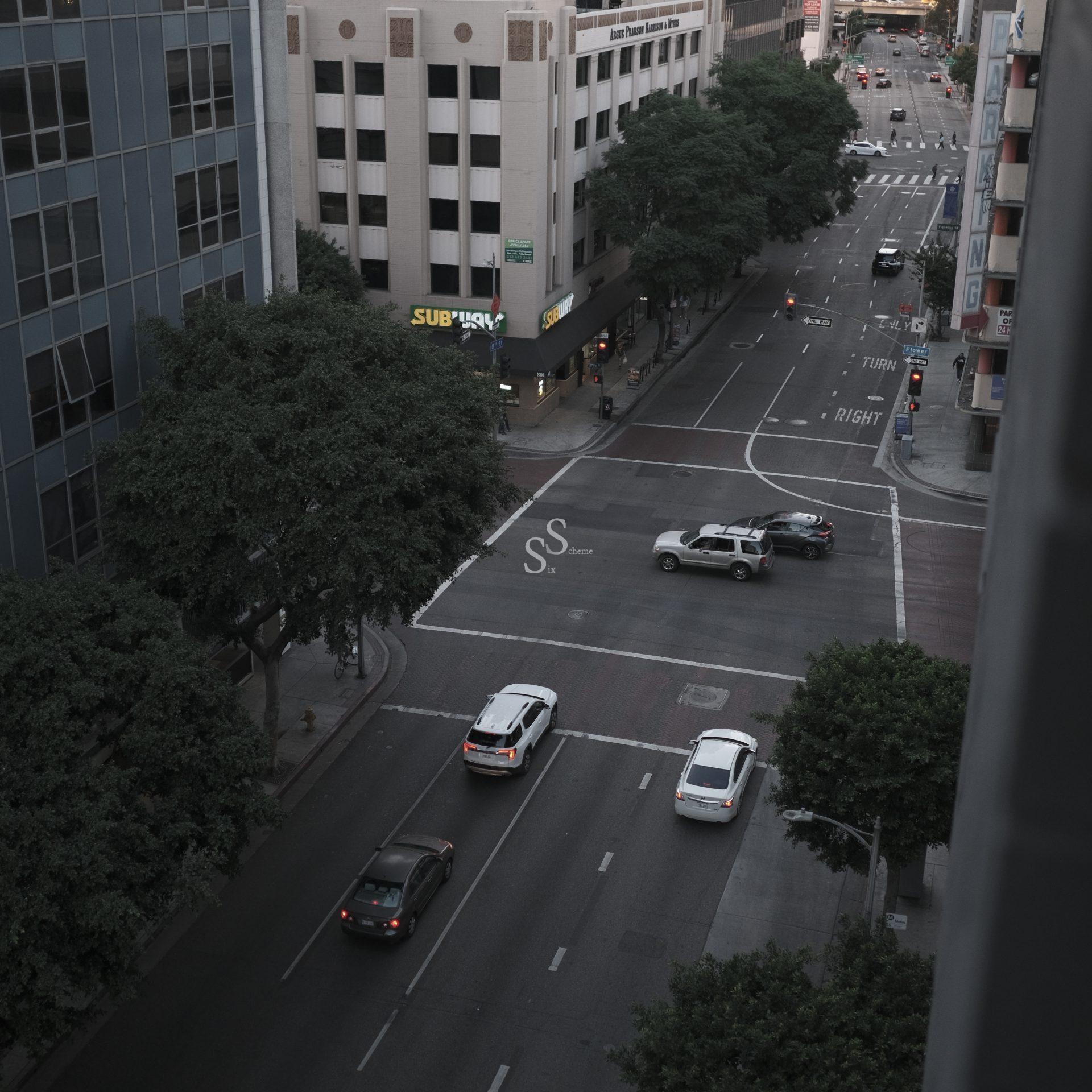 A city street intersection seen from above, with cars stopped at a traffic light. Tall buildings and trees line the roads. A SUBWAY restaurant sign is visible on a corner. The streets appear quiet and mostly empty.