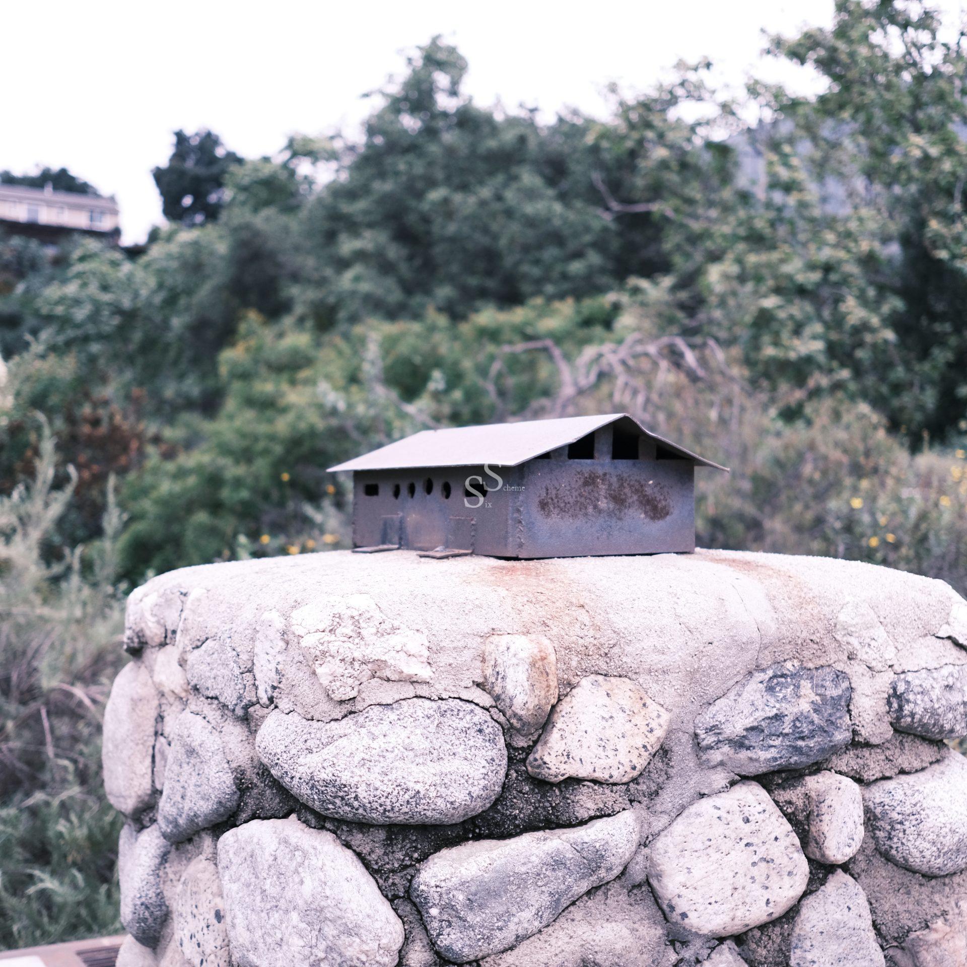 A small metal model of a building sits on top of a round stone pedestal, surrounded by greenery and trees in the background.