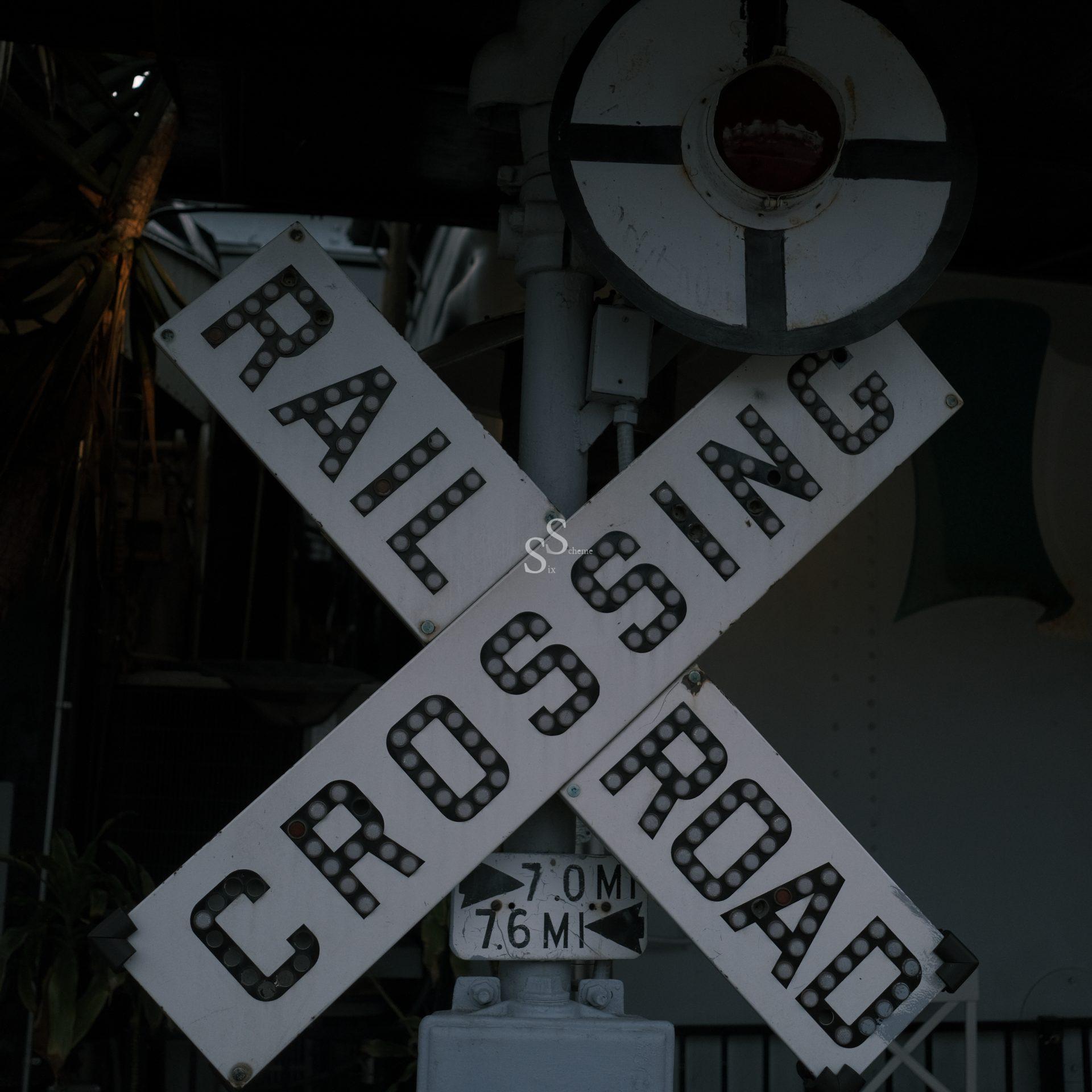 A close-up of a weathered railroad crossing sign with black letters reading RAILROAD CROSSING in an X shape, mounted on a pole. The background is dimly lit and partially obscured.