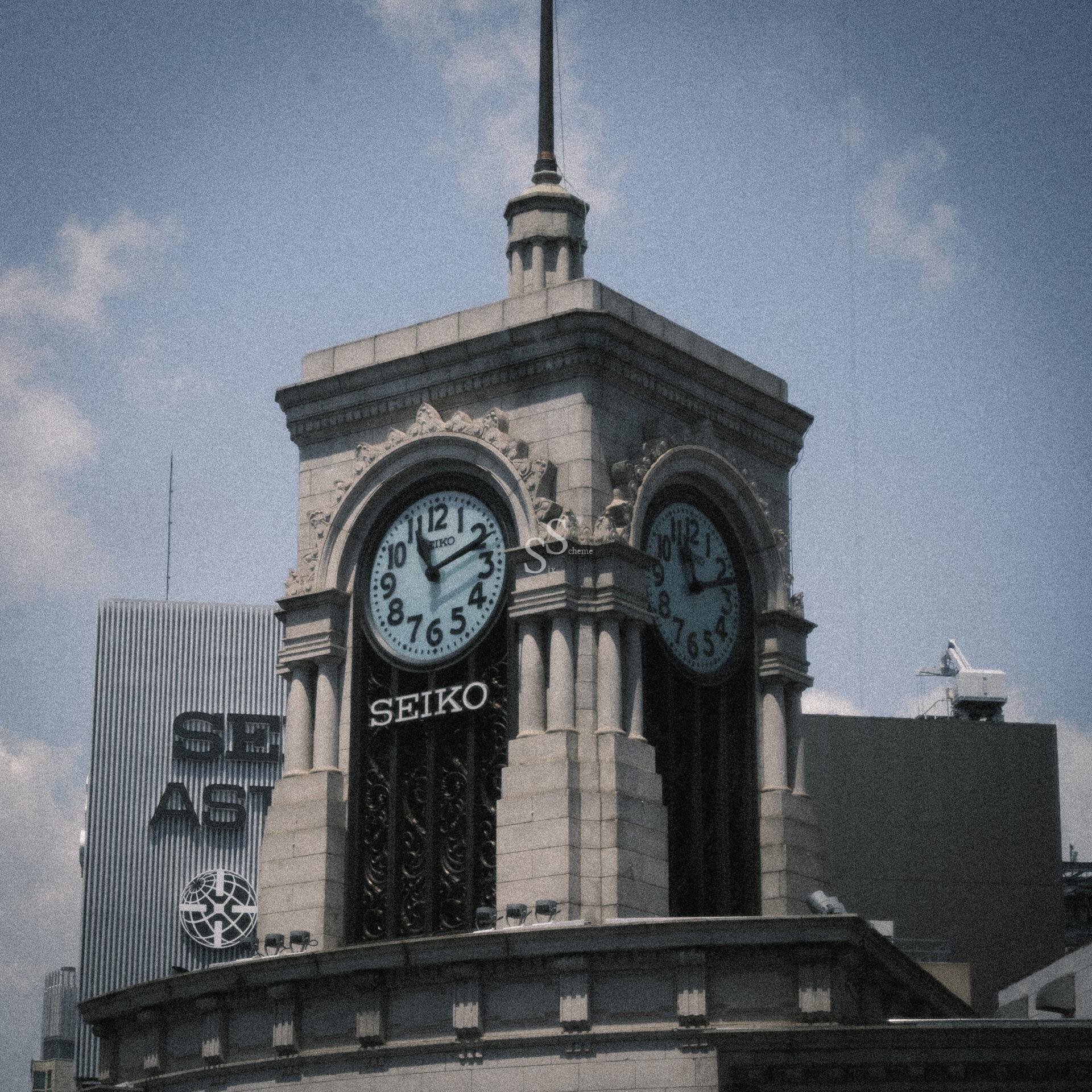 A stone clock tower with two clock faces showing different times stands against a partly cloudy sky. The building features columns and the word SEIKO below the clocks. Nearby modern buildings are visible in the background.