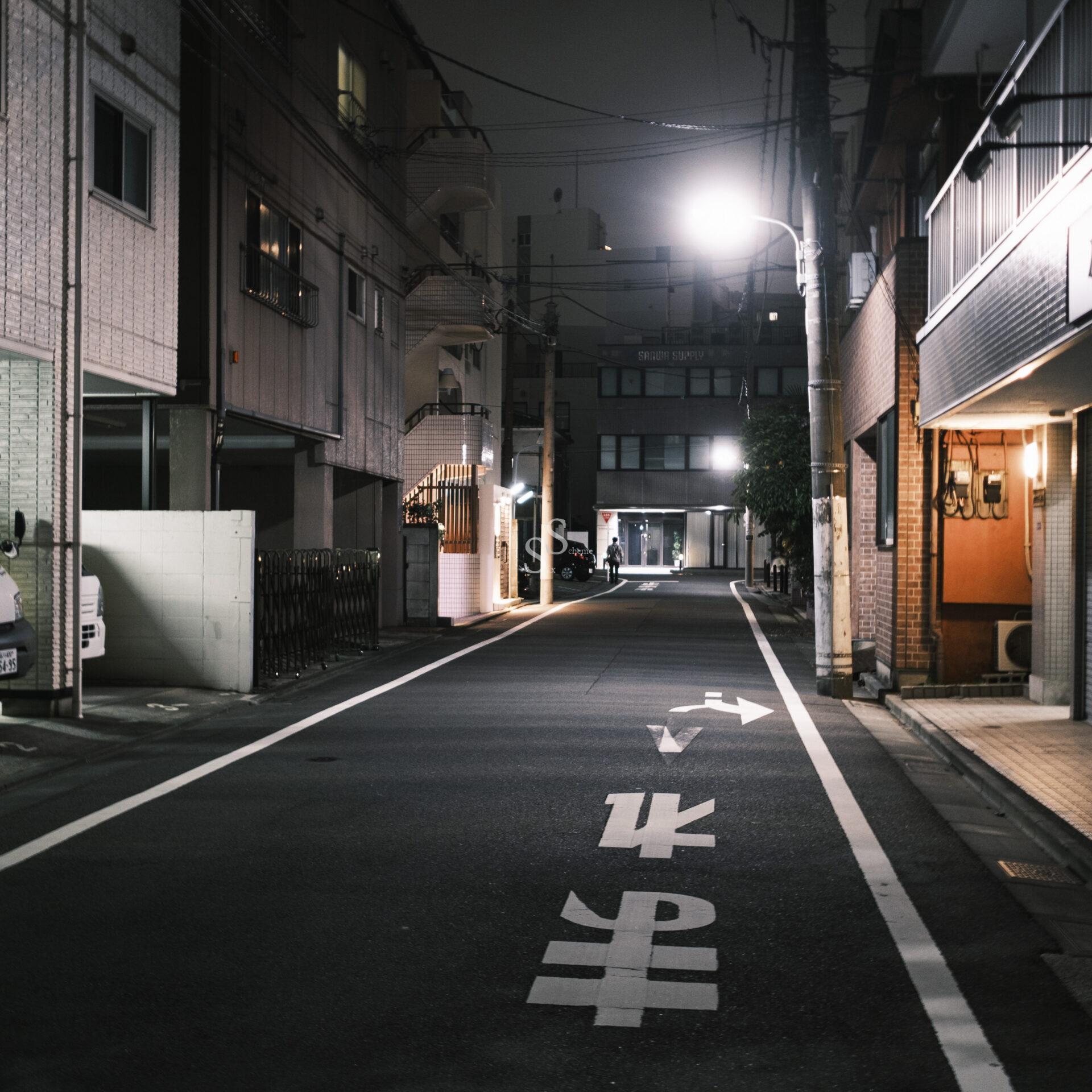 A quiet, empty street at night in a Japanese city, lit by streetlights. Buildings line both sides, and Japanese characters are painted on the road, with no people or cars in sight.