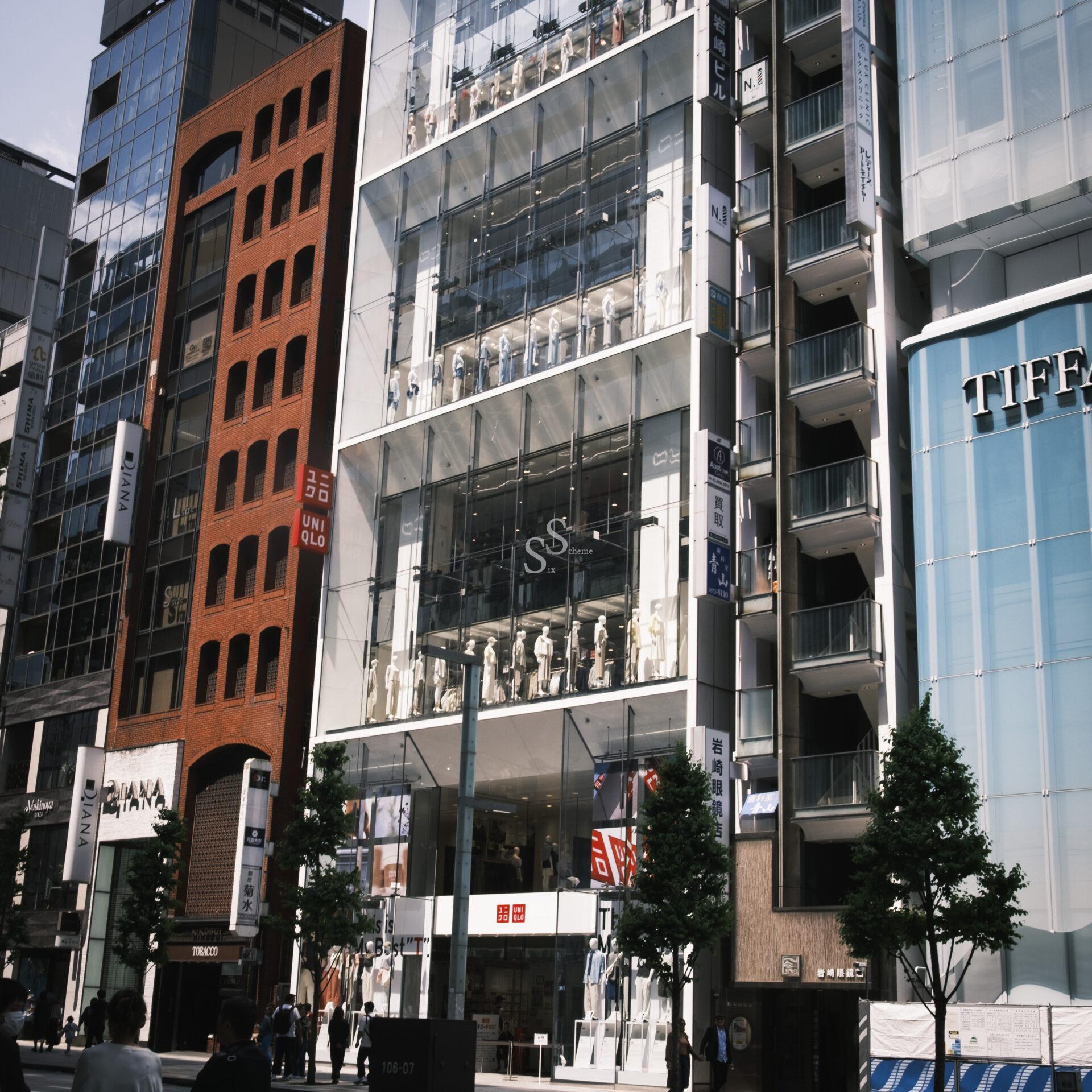 A multi-story Uniqlo store with glass windows displays mannequins in various outfits. The building is surrounded by other modern buildings on a busy city street with people and trees in front.