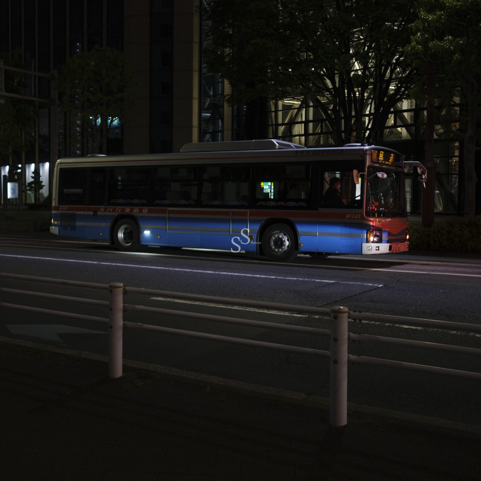 A city bus with blue and red stripes is stopped on a street at night, illuminated by streetlights. The background is dark, with buildings and trees faintly visible.