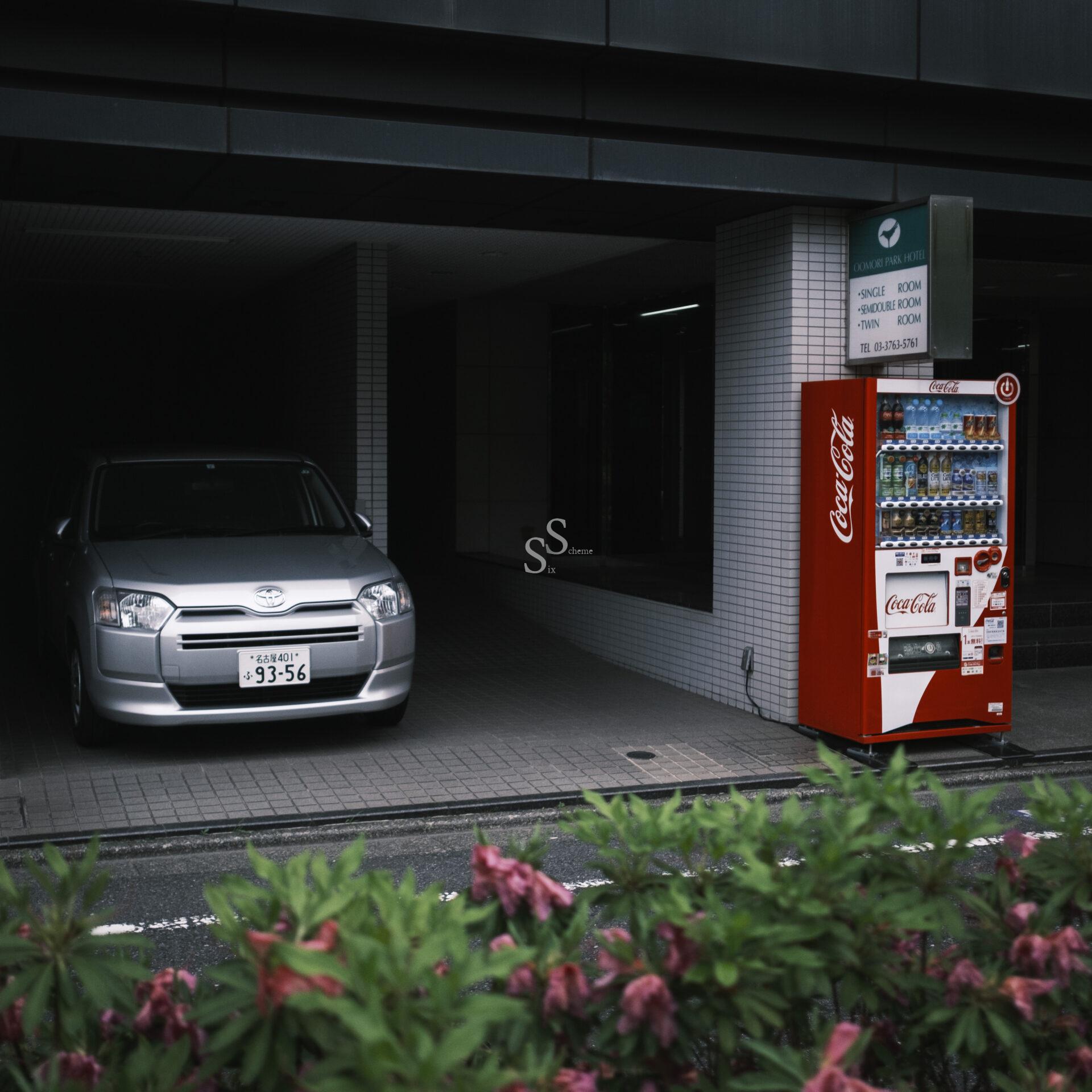 A silver car is parked in a dark building entrance next to a red Coca-Cola vending machine on a sidewalk, with green bushes and pink flowers in the foreground.