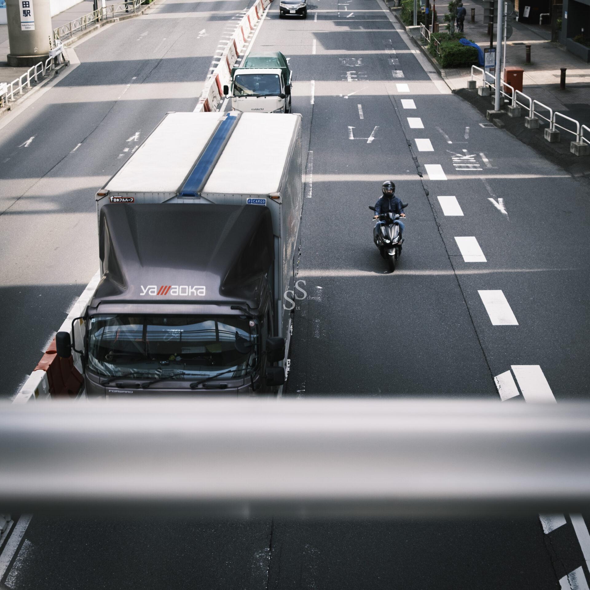 A truck, a car, and a motorcycle travel on a multi-lane city street, with the truck in the left lane and the motorcycle in the right. The photo is taken from above, with a white railing partially visible in the foreground.