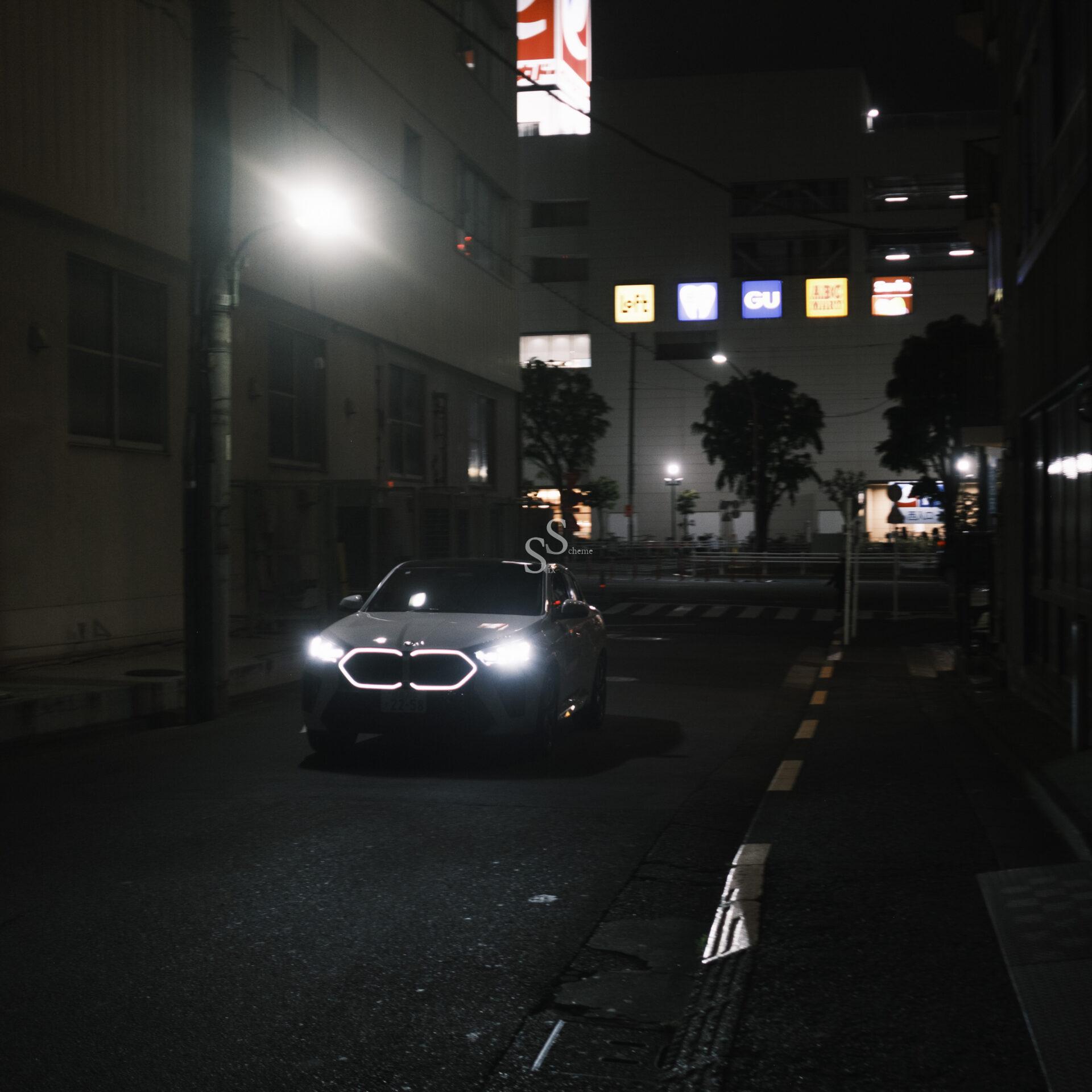 A car with illuminated headlights drives down a dark, empty street at night in a city, with glowing store signs and building lights in the background.