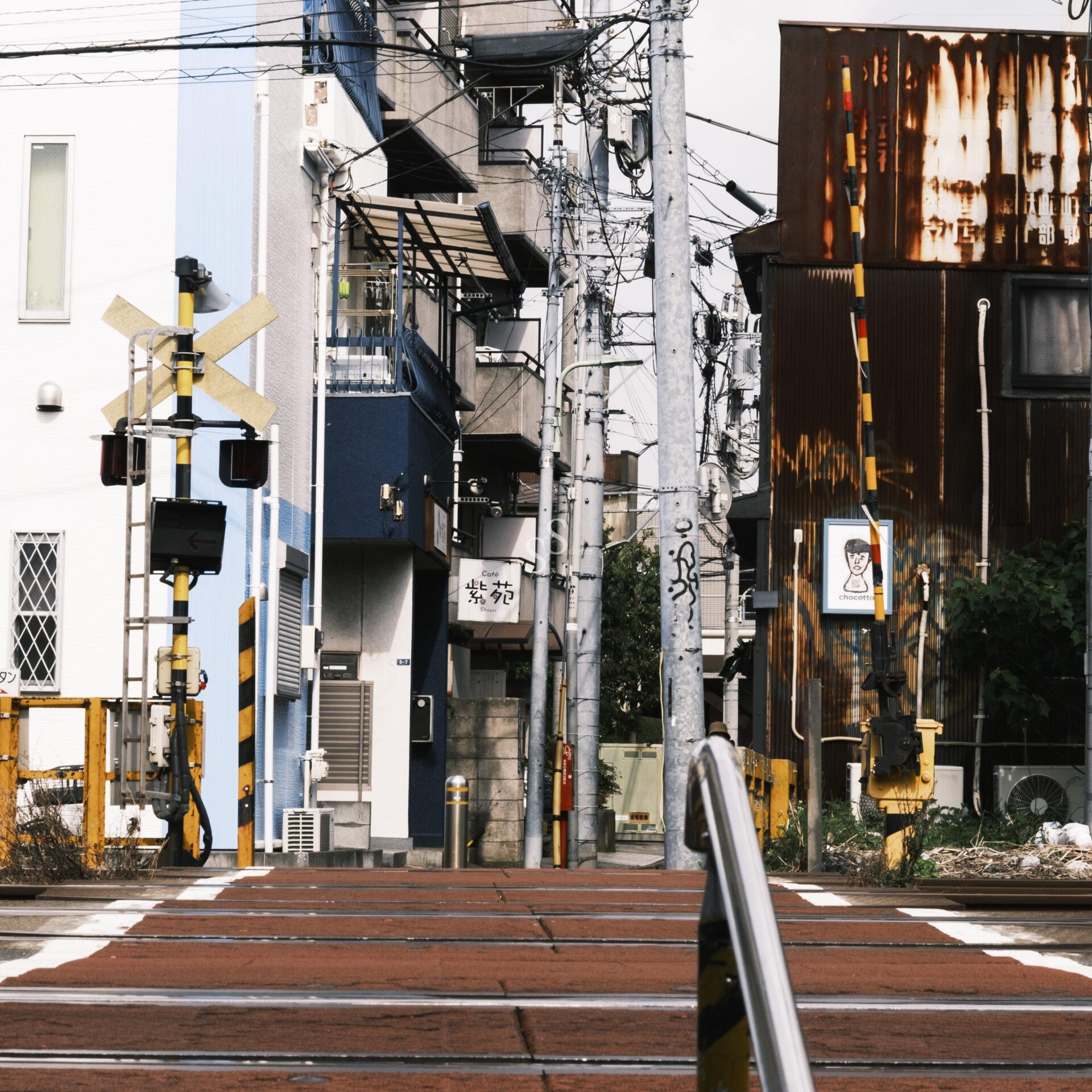 A railway crossing in a city with yellow and black striped barriers, overhead wires, and buildings on both sides, including a rusted metal building and a blue and white one. The crossing gate is up.