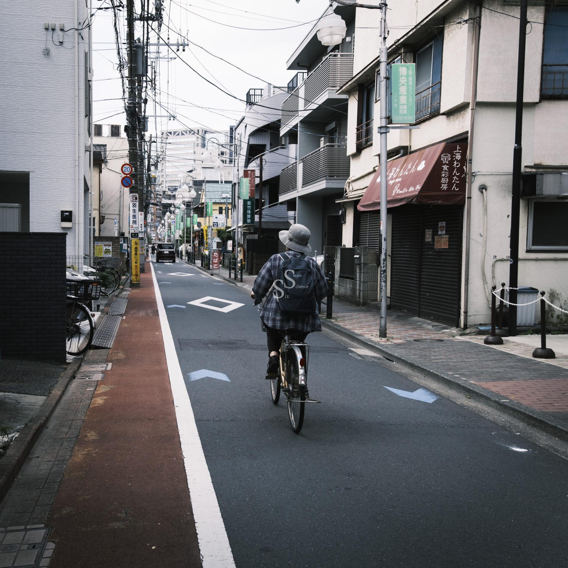 A person wearing a backpack and a bucket hat rides a bicycle down a quiet city street lined with buildings, stores, and power lines on an overcast day.