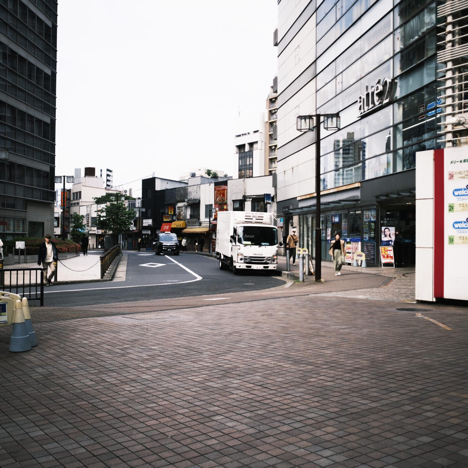 A city street scene with a white truck driving by modern glass buildings, pedestrians walking on the sidewalk, and stores visible. The area is paved with tiles and appears clean and bright.