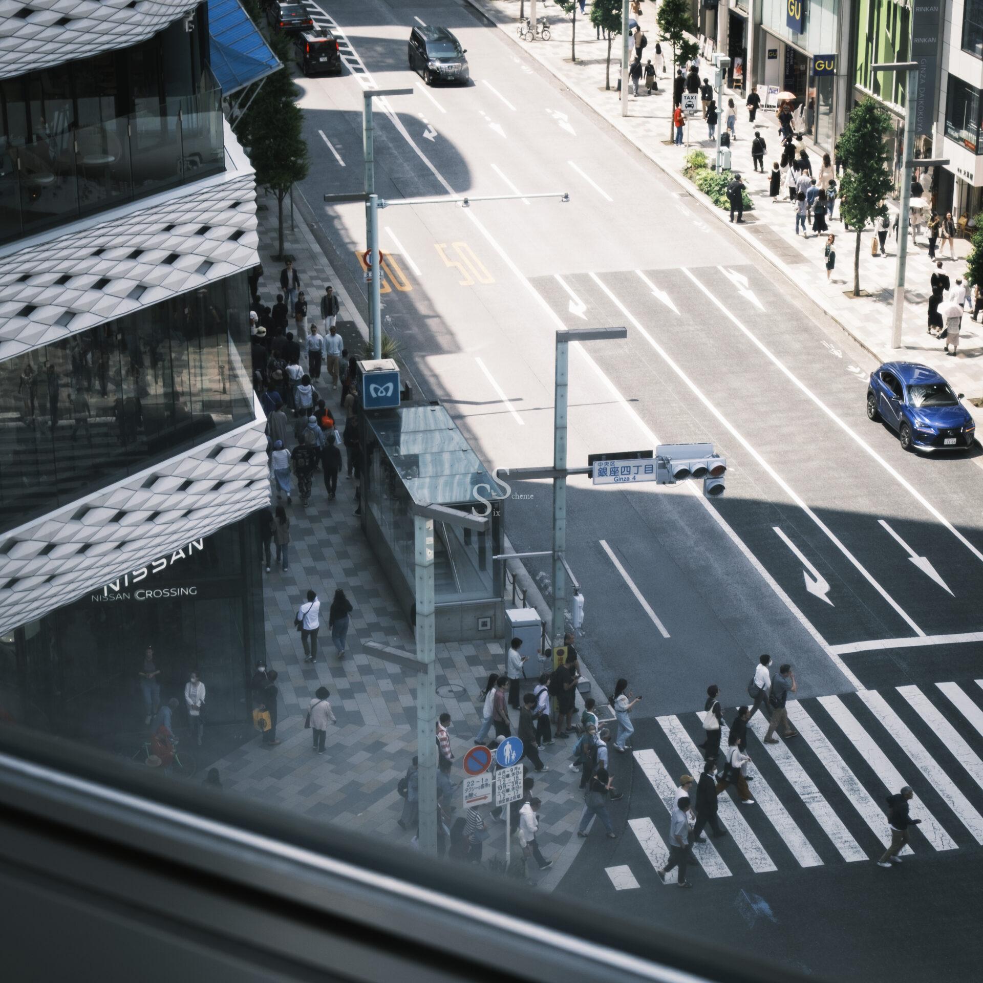 A busy city street seen from above, with people crossing at a crosswalk, cars driving by, and tall buildings lining both sides of the road on a sunny day.