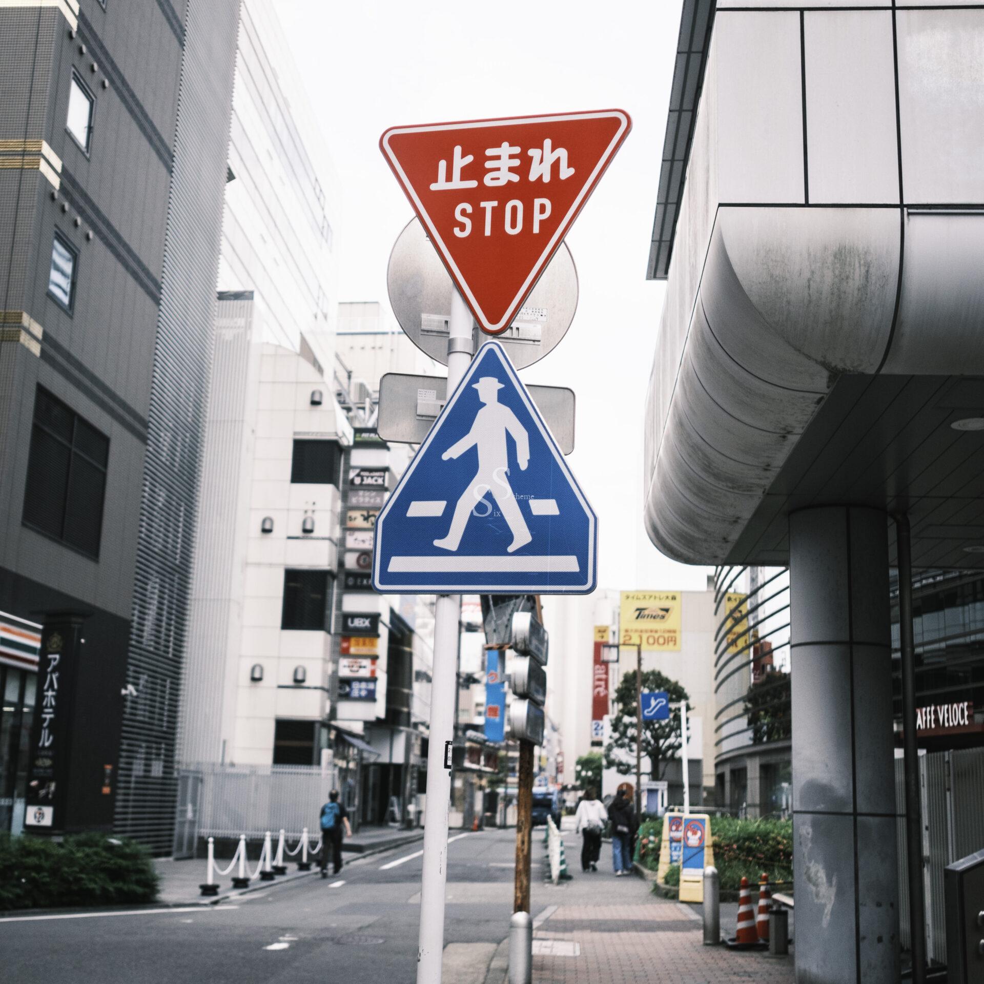 A red stop sign with Japanese and English text is above a blue pedestrian crossing sign on a city street lined with modern buildings and a few people walking.
