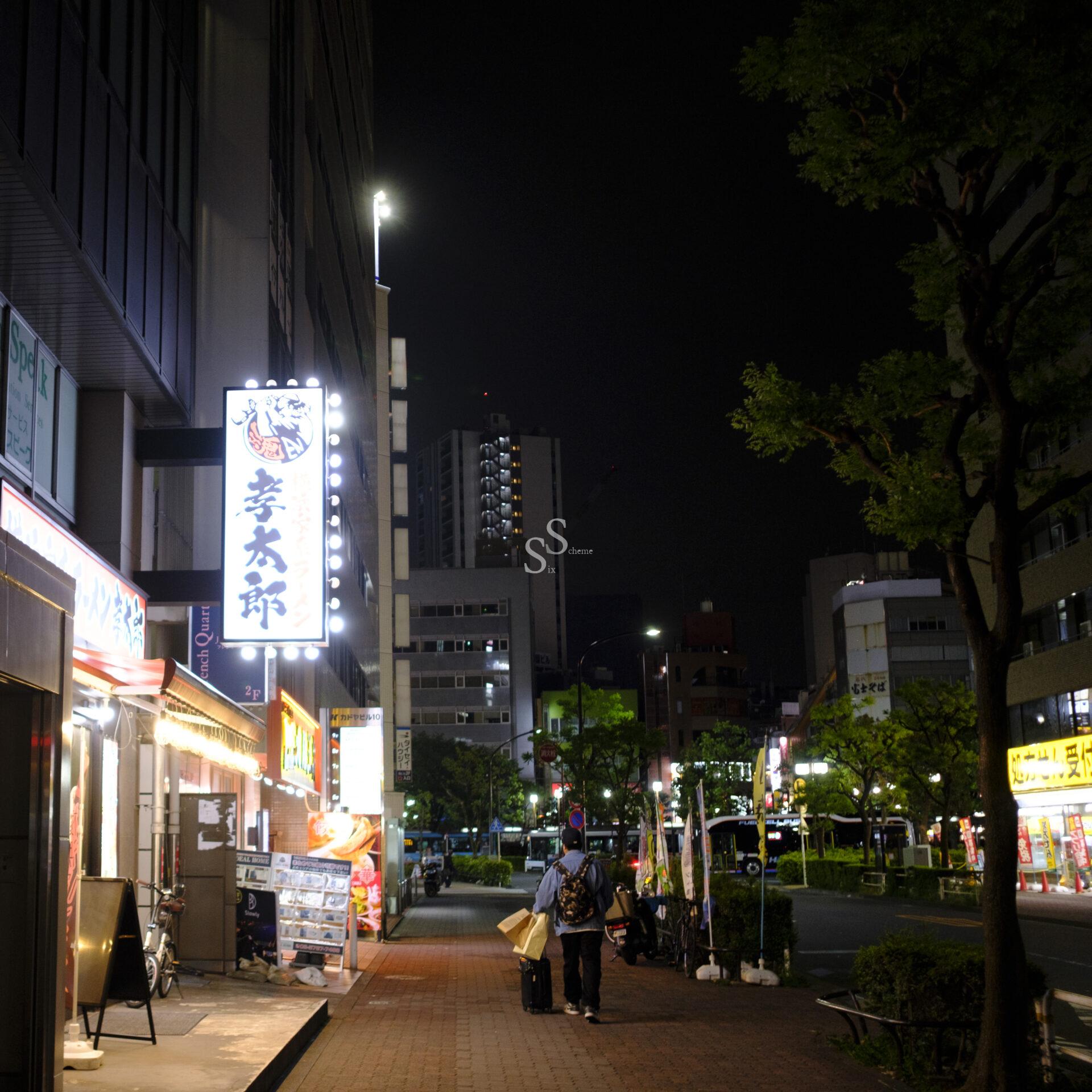 A city street at night with neon signs, illuminated shopfronts, and a few people walking on the sidewalk. Tall buildings and trees line the street, and the sky is dark.