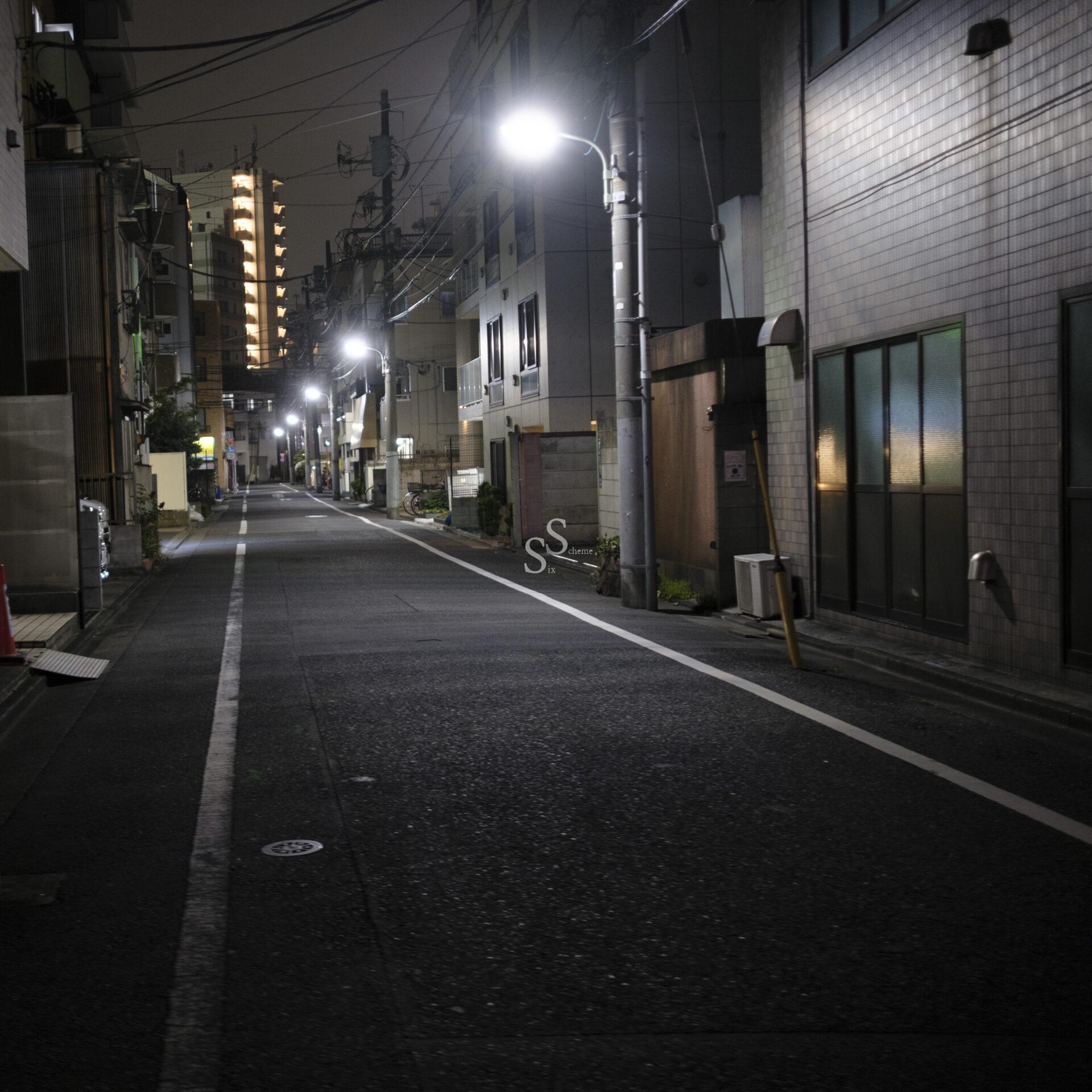 A quiet, empty street at night in an urban area, illuminated by streetlights. Tall buildings line both sides, and windows glow softly. The scene feels calm and deserted.