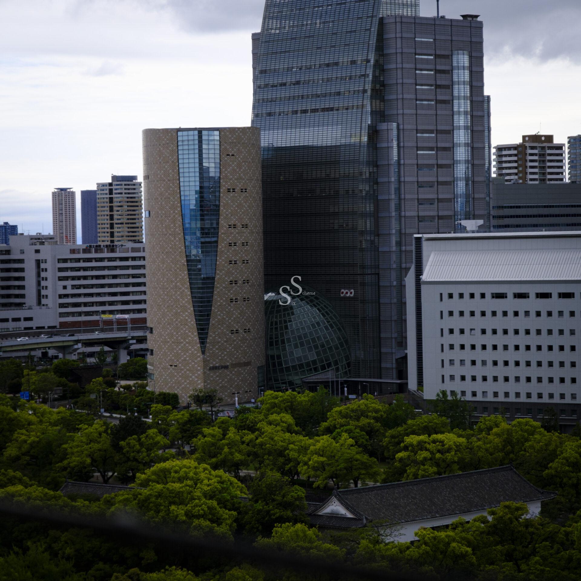 A cityscape featuring modern high-rise buildings with glass and textured facades, surrounded by lush green trees in the foreground under a cloudy sky.