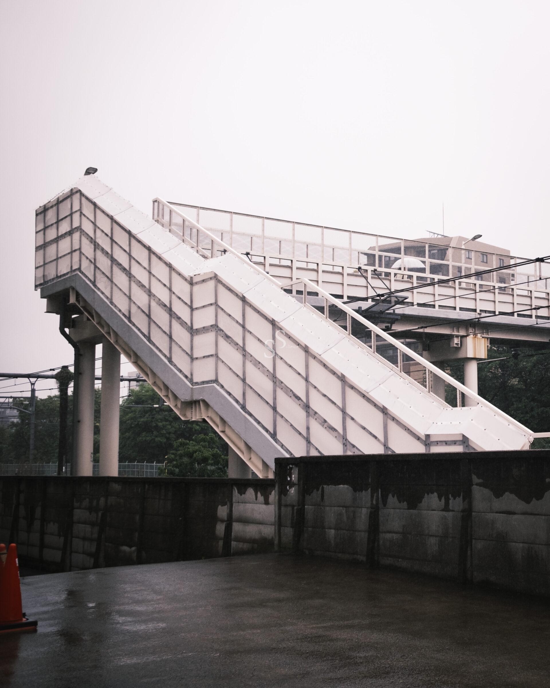 A covered pedestrian bridge with white panels and railings crosses over a wet, empty concrete area on a cloudy day. Two orange traffic cones are visible in the foreground. Trees are in the background.