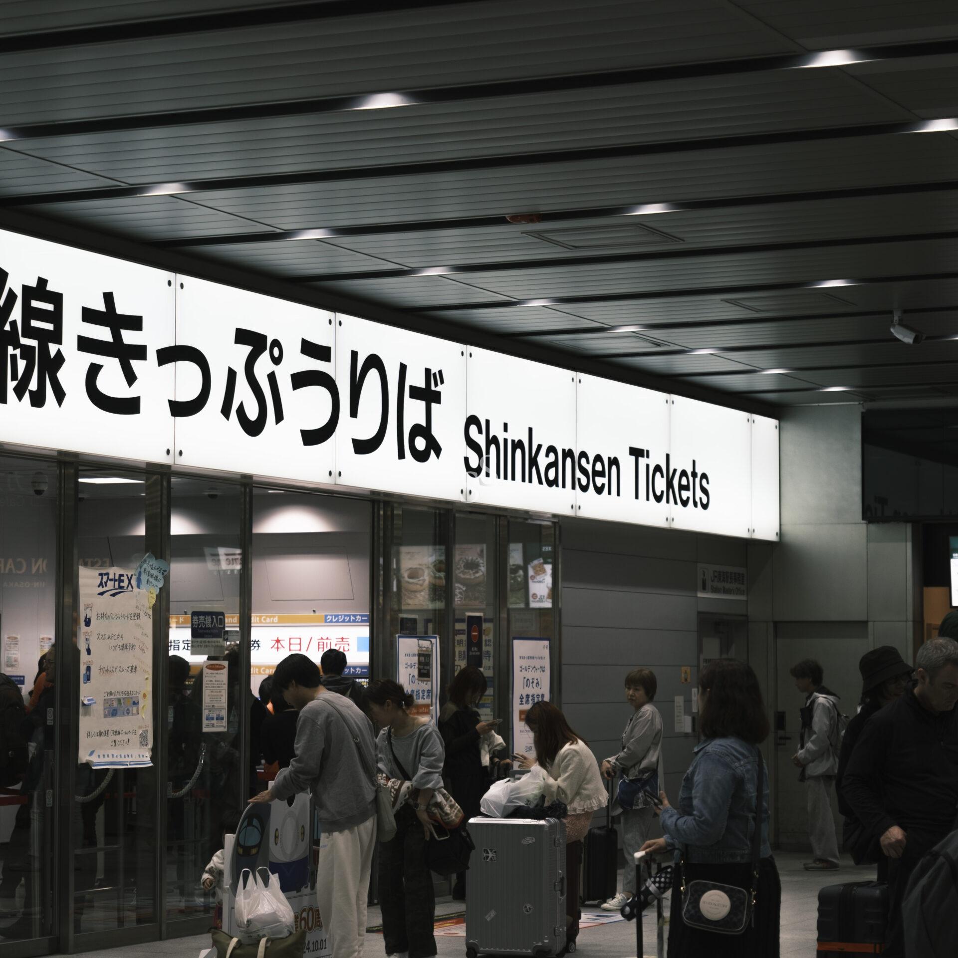 People wait in line at a Shinkansen ticket counter inside a train station in Japan. A large bilingual sign above reads “Shinkansen Tickets” in both Japanese and English.