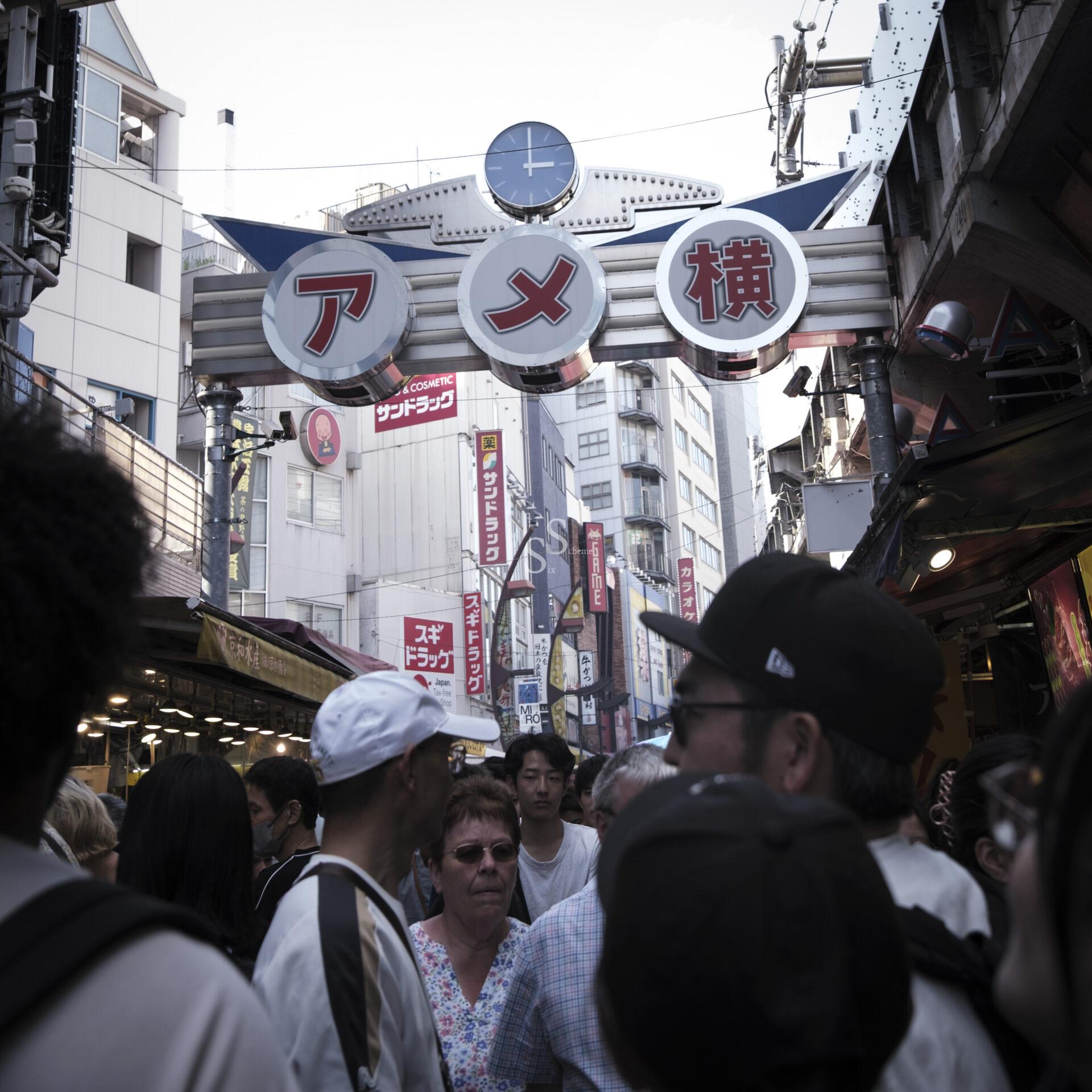 A crowded street market in Japan with a large sign overhead displaying Japanese characters and a clock. People fill the bustling walkway, surrounded by shops and tall buildings.