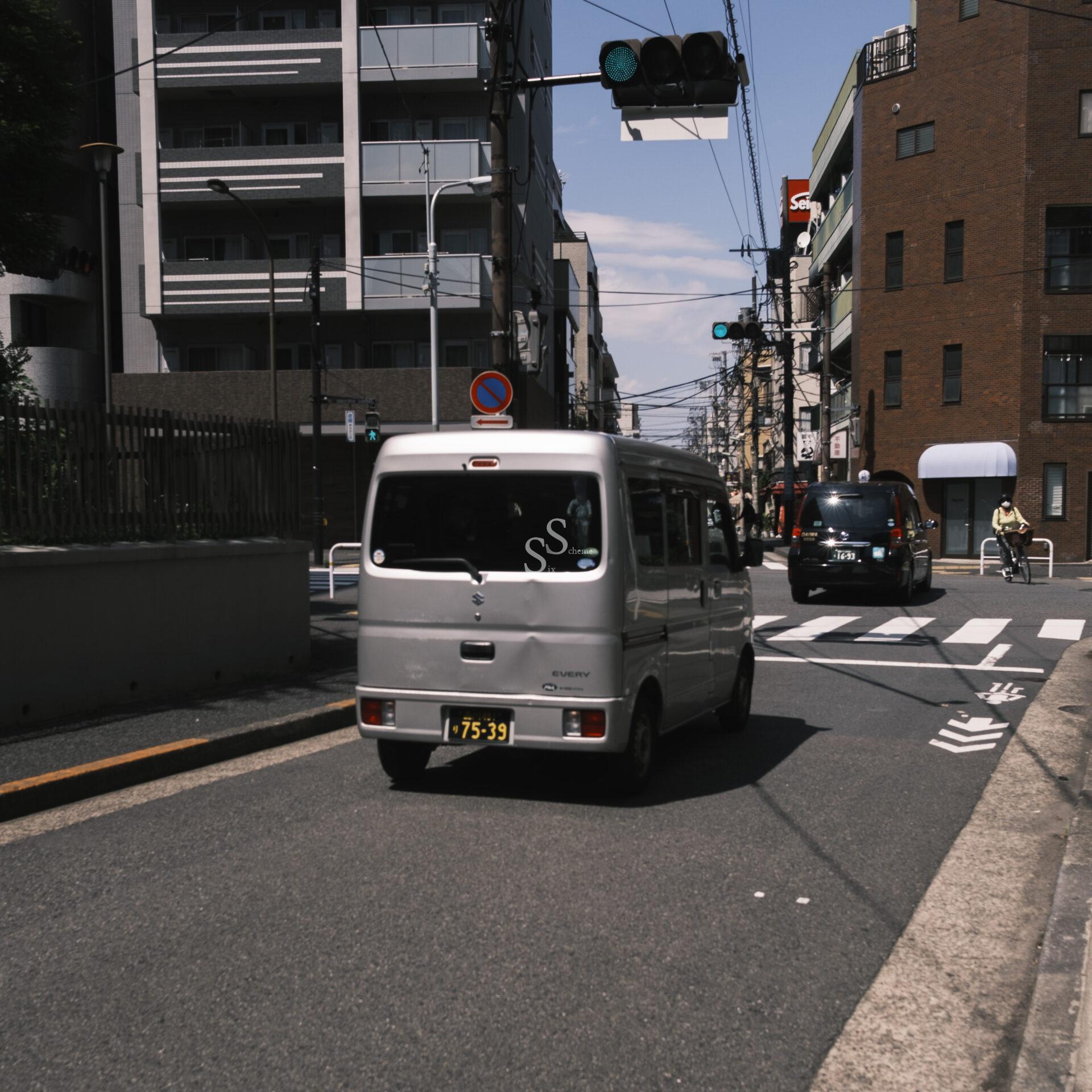 A silver van drives down a city street lined with apartment buildings on a sunny day. A few cars, a cyclist, and various street signs are visible in the background.