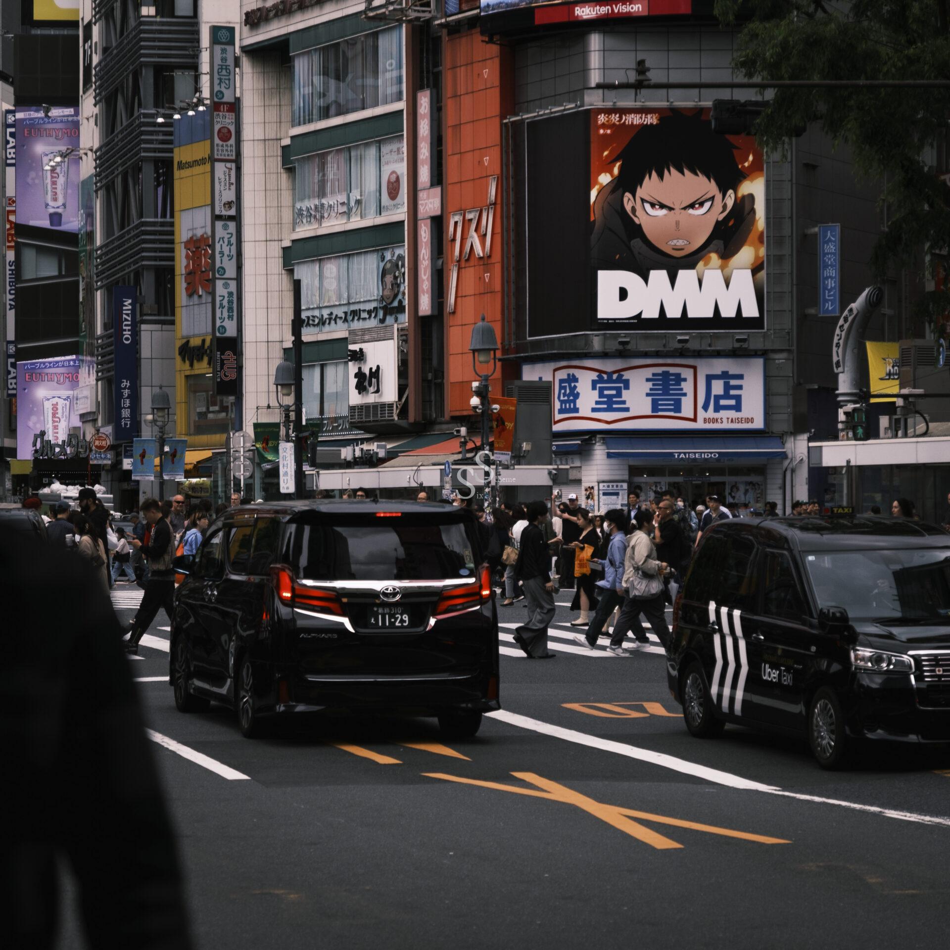 Busy city intersection with crowds crossing the street, black cars driving by, and tall buildings with colorful signs. A large billboard displays an anime character above the scene.