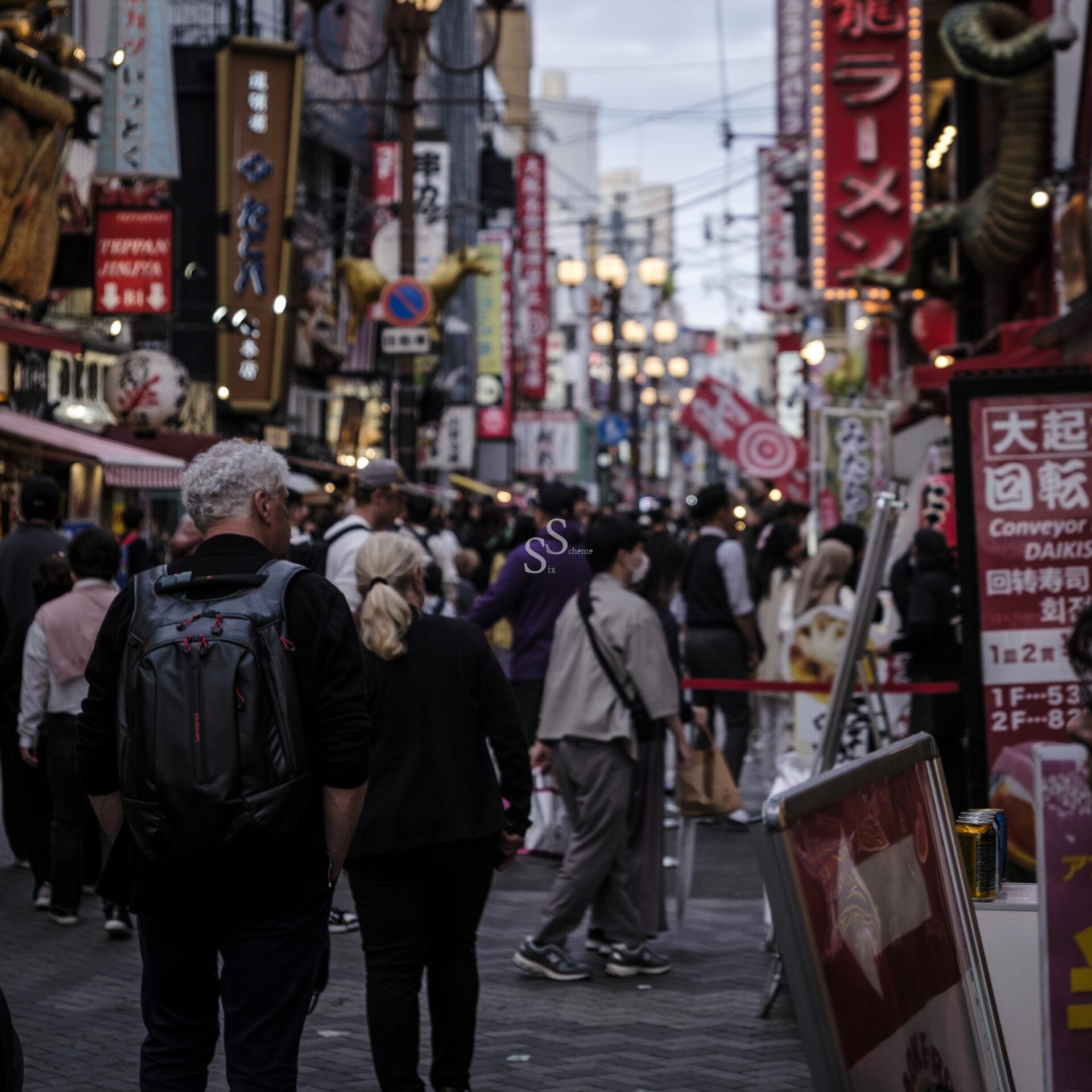 A crowded street in Japan lined with colorful signs, lanterns, and restaurants. People walk along the busy path, some wearing backpacks and masks, as evening light fills the vibrant urban scene.