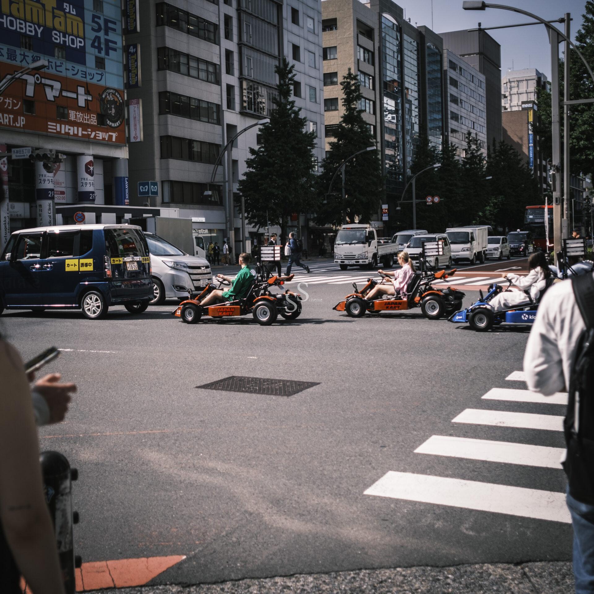 People in costumes drive go-karts on a city street alongside cars, with tall buildings, shops, and a crosswalk in the background. Pedestrians are visible in the foreground.