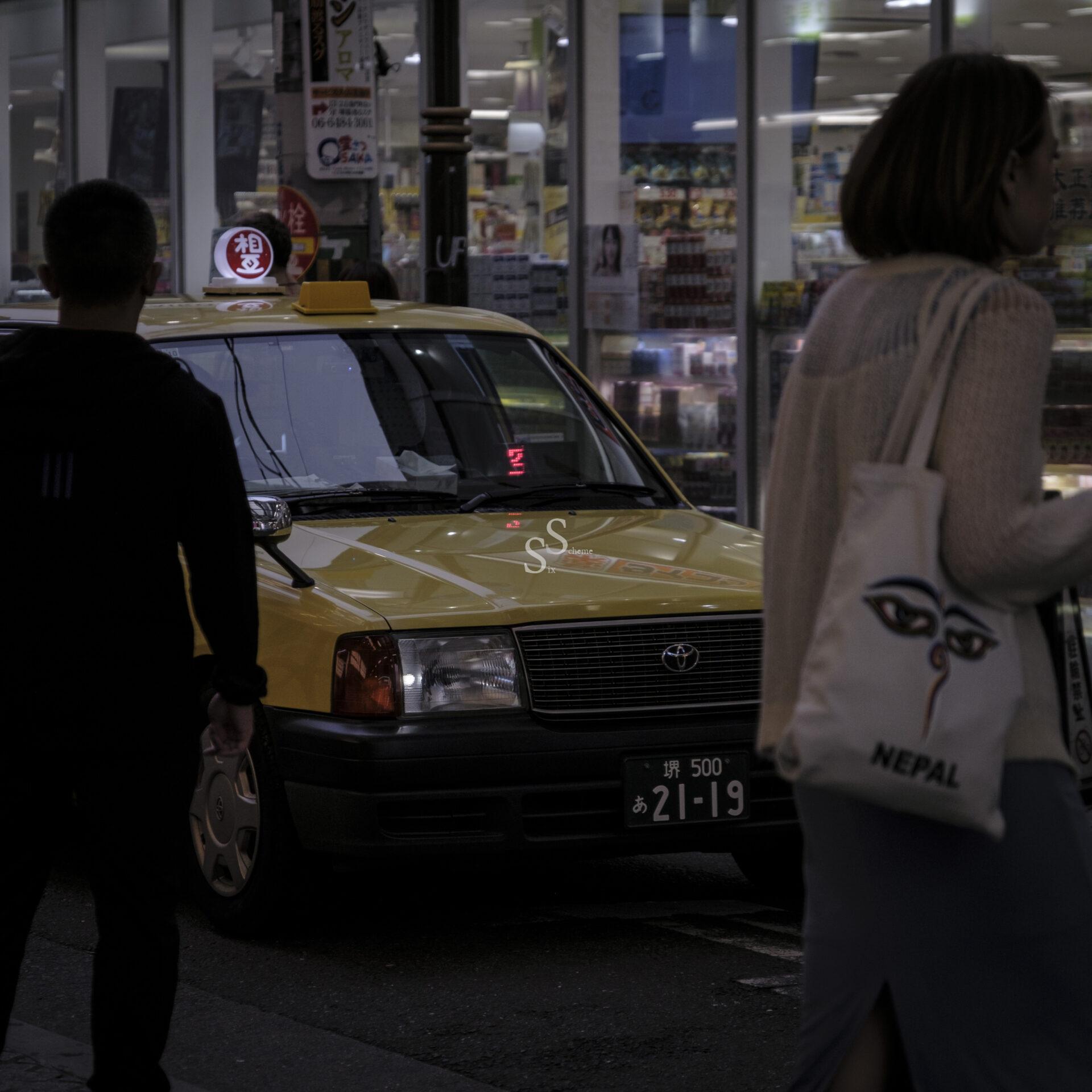 A yellow taxi parked on a dimly lit street at night, with two people walking past; one carries a white tote bag with NEPAL and an illustrated eye printed on it. Storefront lights are visible in the background.