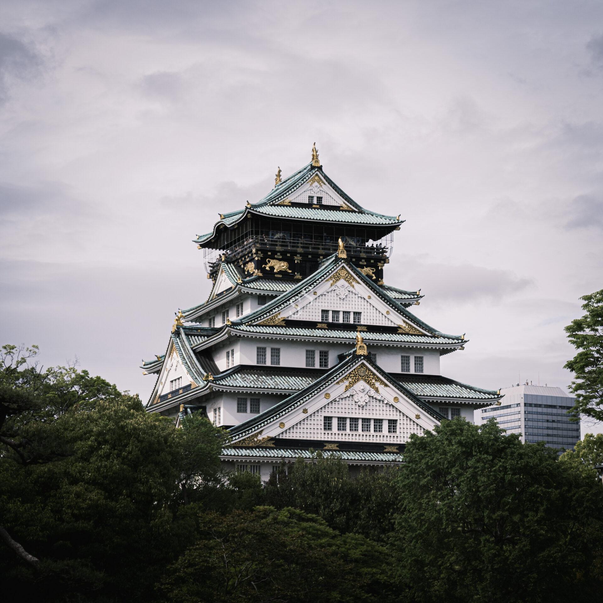Osaka Castle with its white walls, green-tiled roofs, and gold accents stands tall amid lush green trees under a cloudy sky. A modern building is visible in the background.