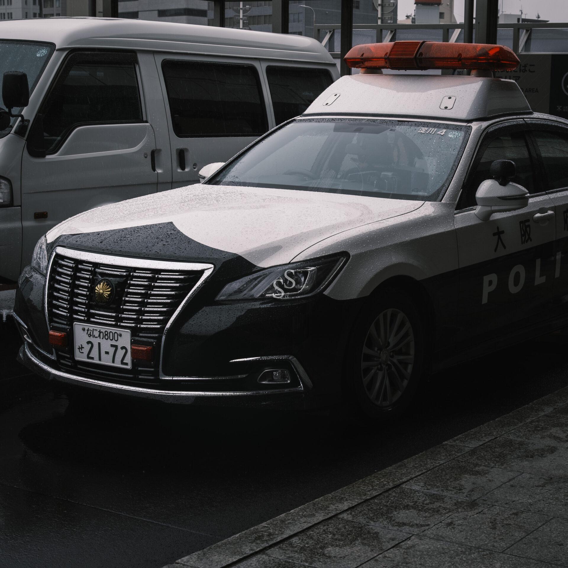 A Japanese police car with a red light bar is parked on a wet street beside a white van. The cars license plate and police markings are visible, and the scene looks overcast or rainy.