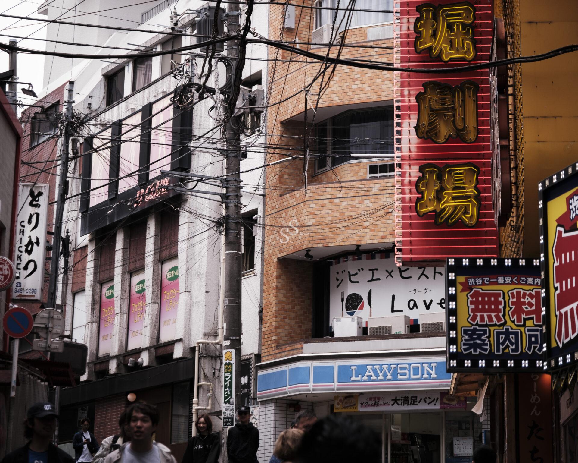 A busy street in Japan with colorful signs, power lines overhead, and people walking. Storefronts include a Lawson convenience store and various signs in Japanese characters. The buildings are close together.