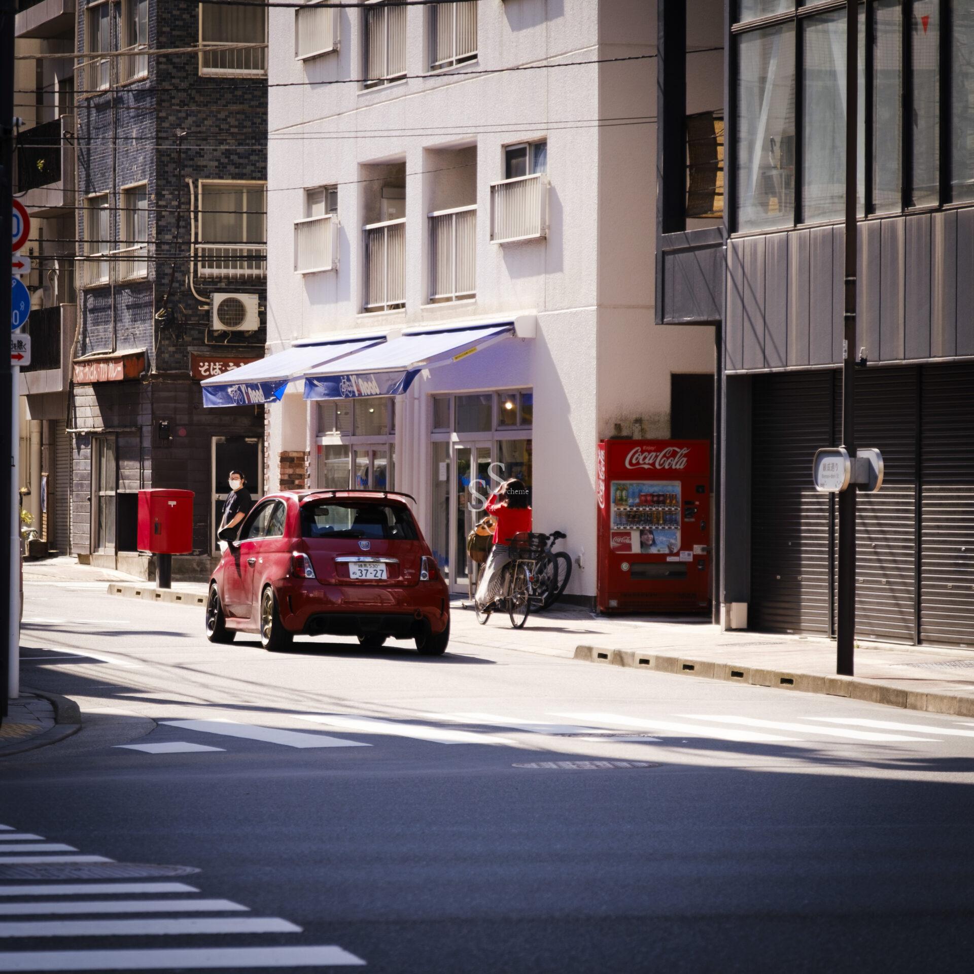 A red car drives down a quiet city street lined with buildings. A person stands by a vending machine next to a bicycle while sunlight casts shadows on the road and sidewalks.