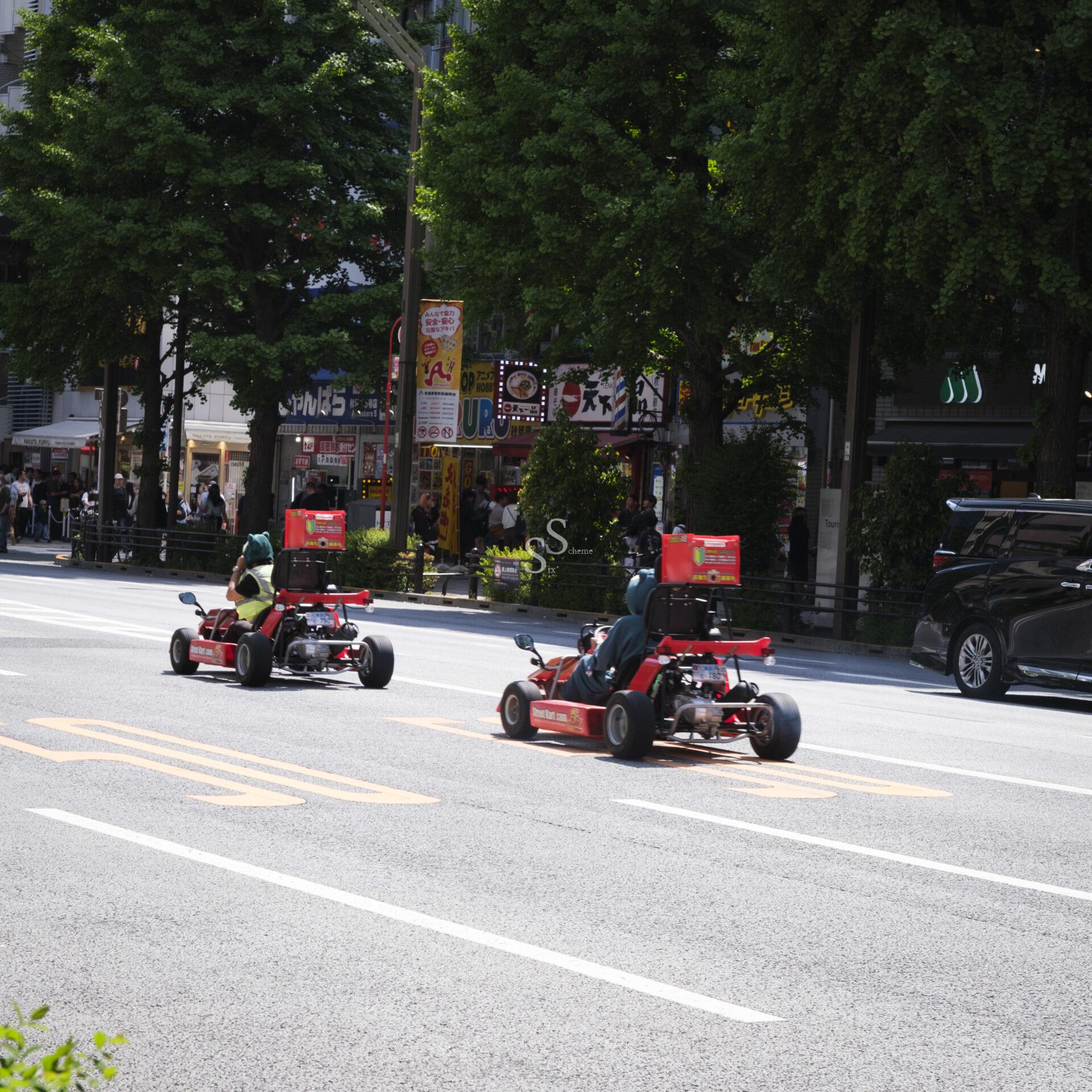 Two people dressed in costumes drive red go-karts on a city street with shops, trees, and parked cars in the background. Pedestrians are visible on the sidewalk.