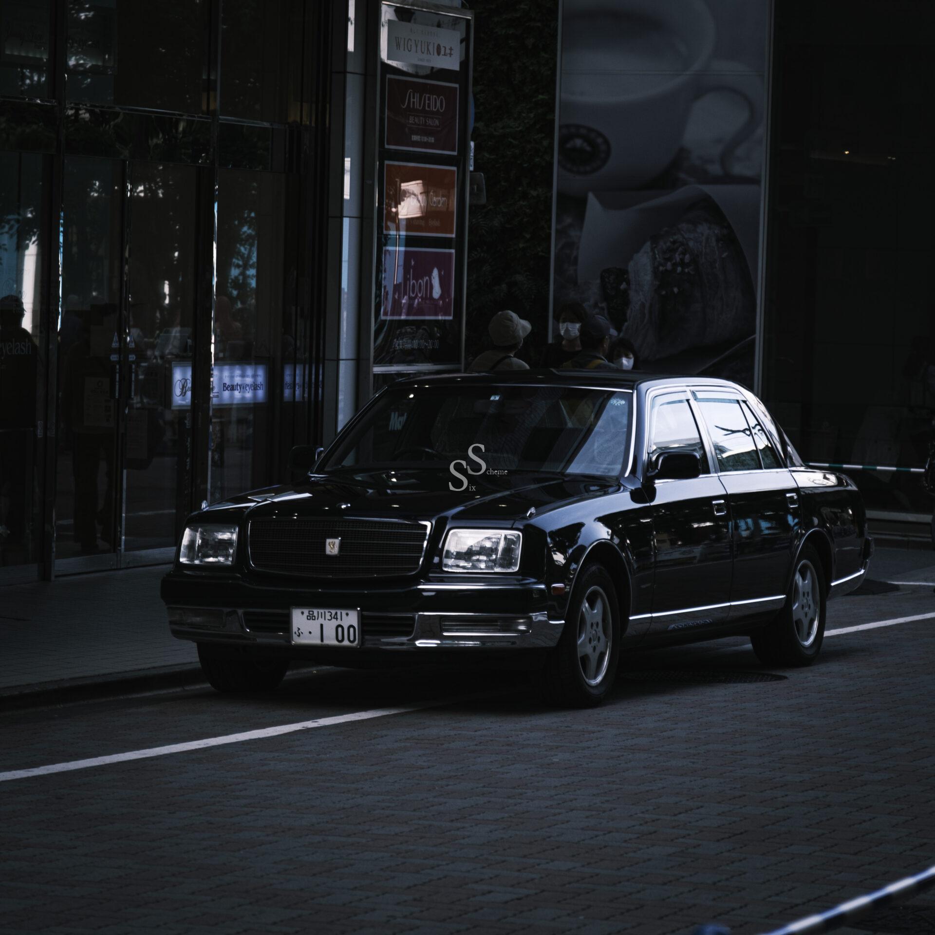 A black luxury sedan with a Japanese license plate is parked on a city street beside a glass building and shop signs, under dim lighting.