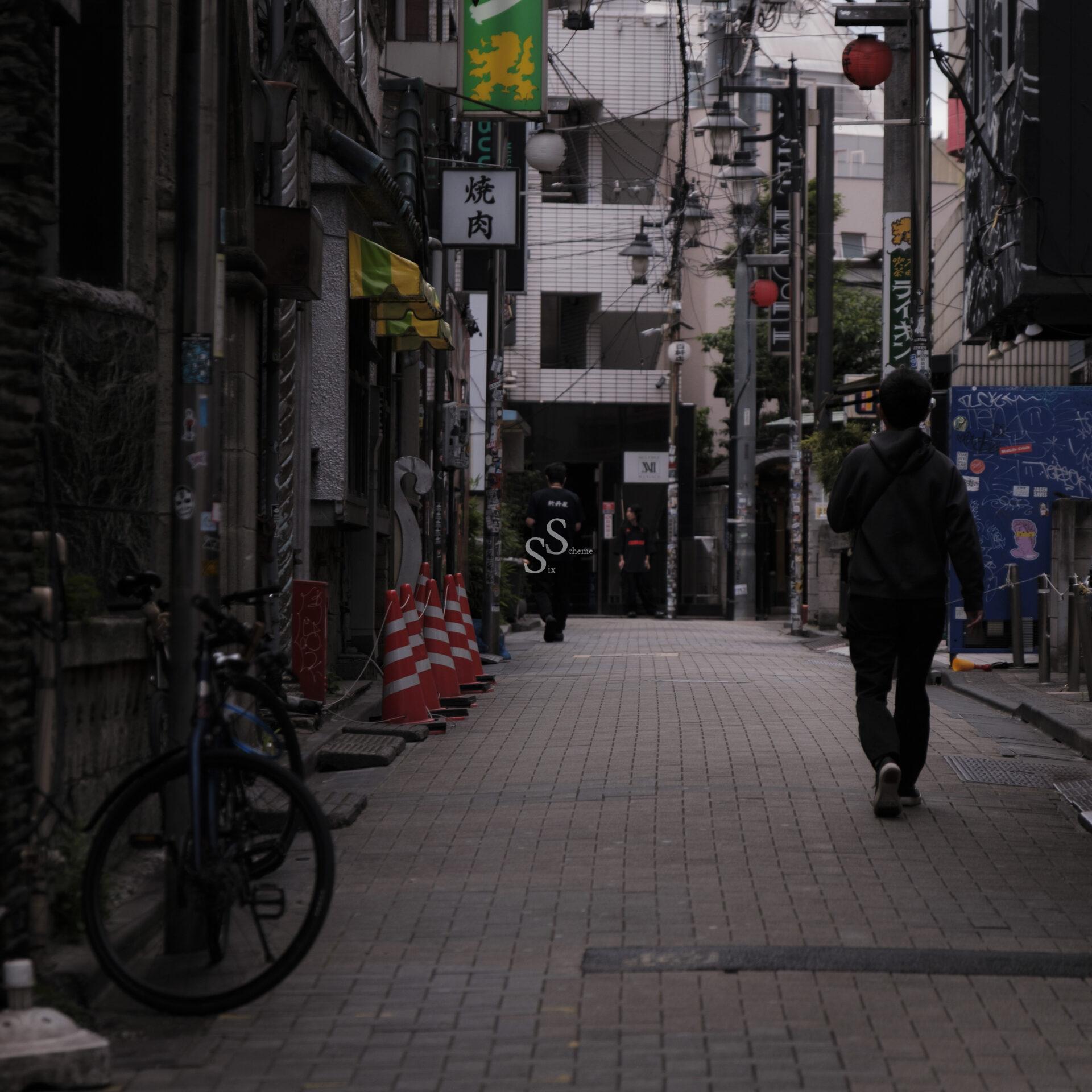 A narrow, dimly lit urban alley with a person walking away, a bicycle parked on the left, orange traffic cones, and various signs and lanterns hanging above the storefronts lining the street.