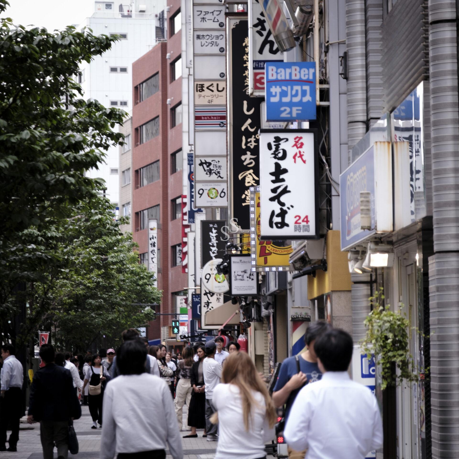 A busy city street in Japan lined with tall buildings, numerous vertical signs in Japanese, and people walking on the sidewalk. Trees line the left side of the walkway. The atmosphere is lively and urban.