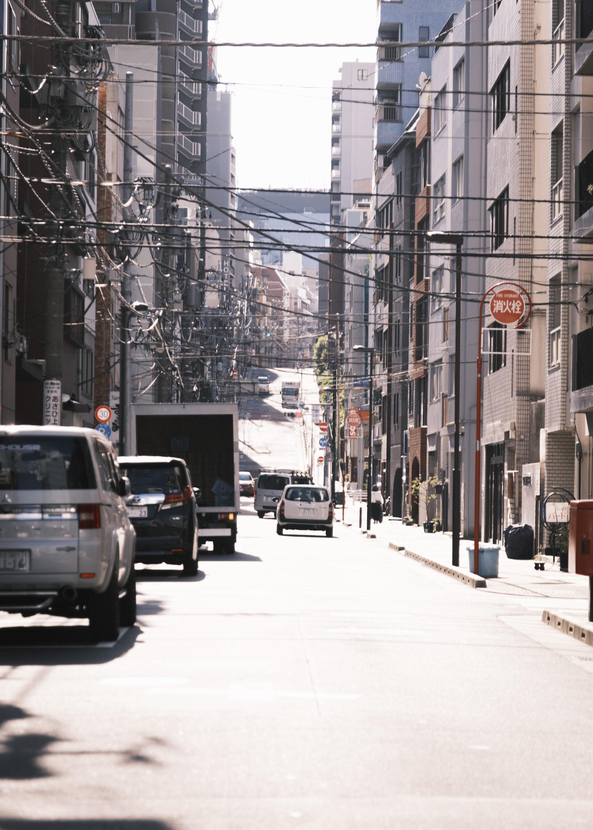 A sunny urban street scene with cars parked and driving, surrounded by tall buildings on both sides and many overhead utility wires. The street slopes upward in the distance.