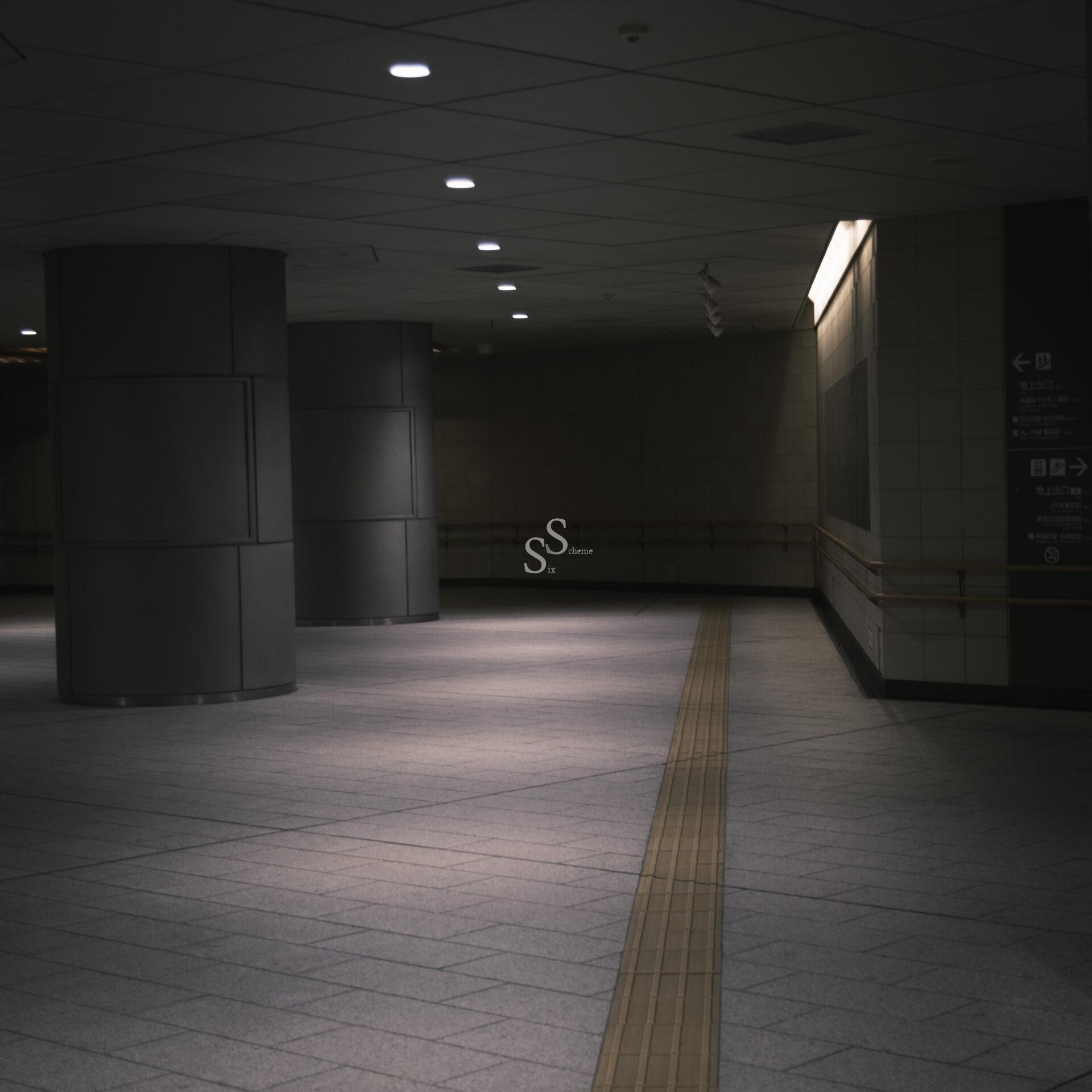 A dimly lit, empty indoor corridor with tiled floors, large dark columns, and tactile paving guiding along the floor for visually impaired individuals. The space has a quiet, modern, and slightly moody atmosphere.