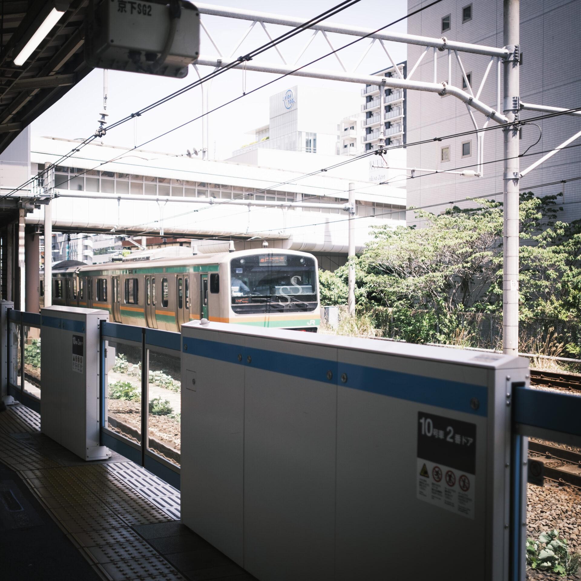 A train arrives at an outdoor urban station with safety barriers. Overhead wires and nearby buildings are visible in the background on a sunny day.