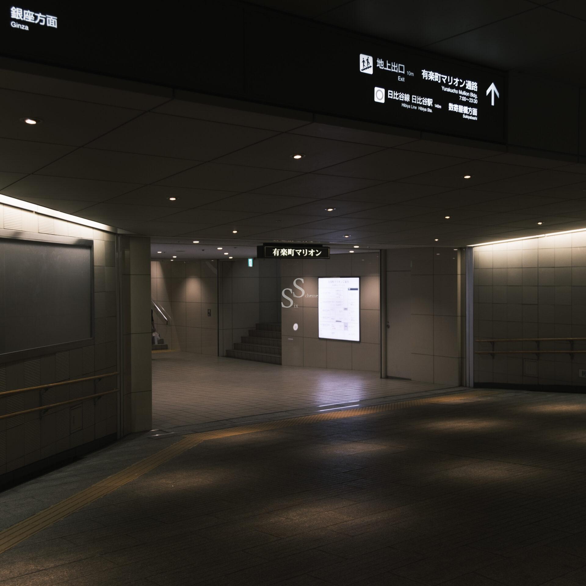 A dimly lit, modern underground passageway in a subway or train station, this liminal space features illuminated signs in Japanese and English, tiled walls, and a ramp leading to an upper level.